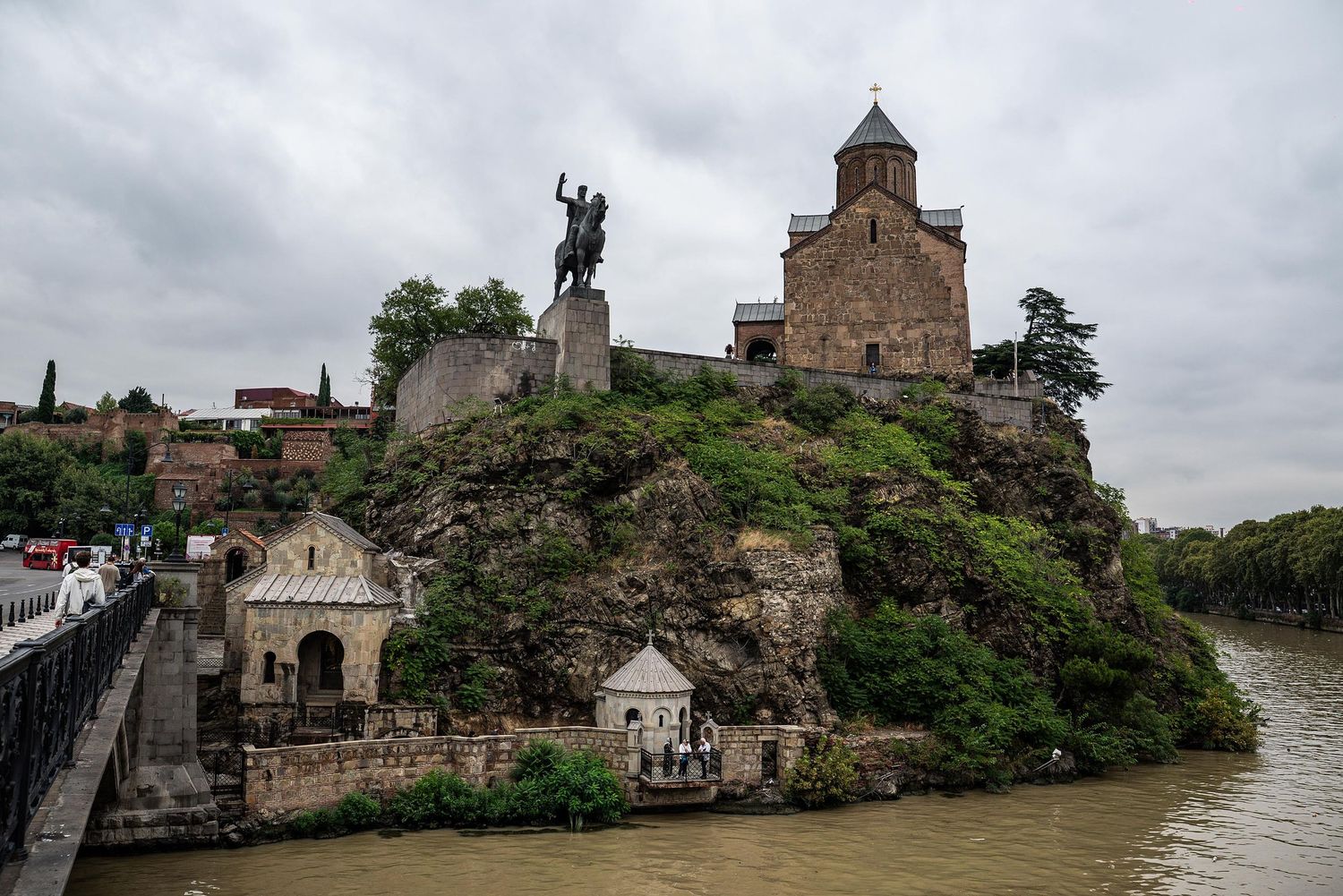 Historic stone buildings and towers line a scenic river bank under cloudy skies in a medieval European setting.