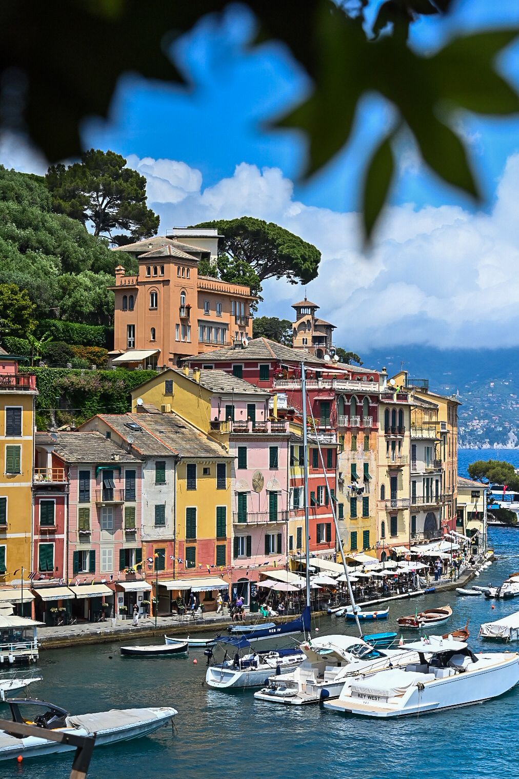 Colorful houses and boats line the waterfront in Portofino, Italy with blue sky and mountains in the background.
