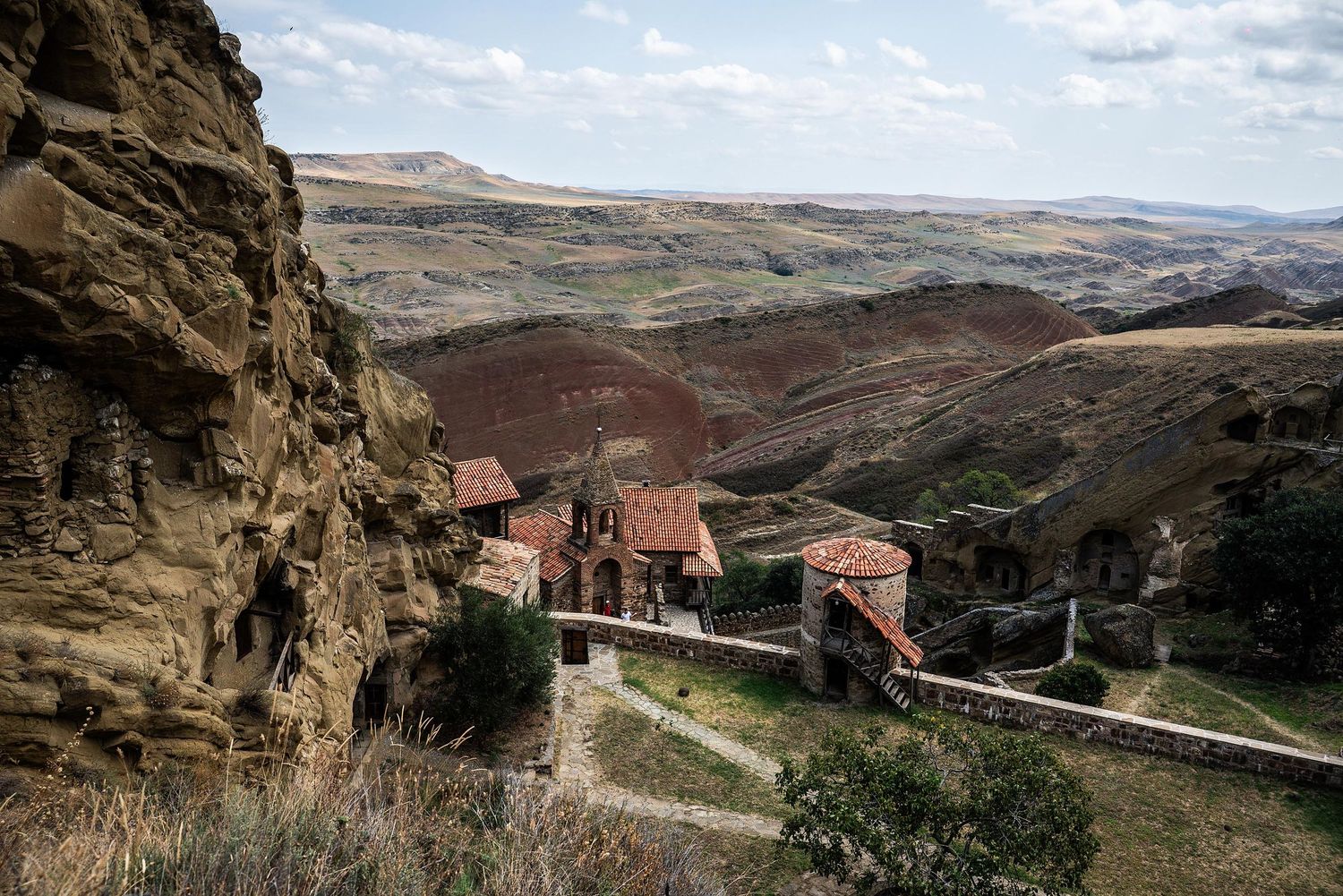 Ancient cave monastery nestled into rugged stone cliffs with panoramic views of the desert landscape and valleys below.