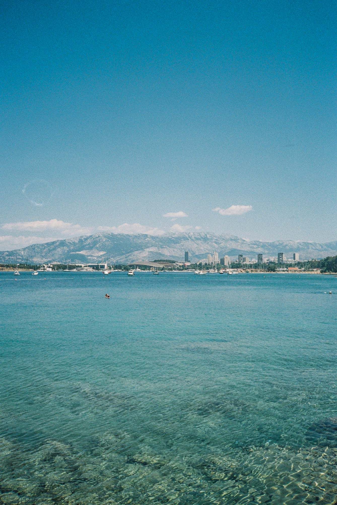 Panoramic view of crystal clear turquoise waters with distant mountains and coastal cityscape under bright blue skies.