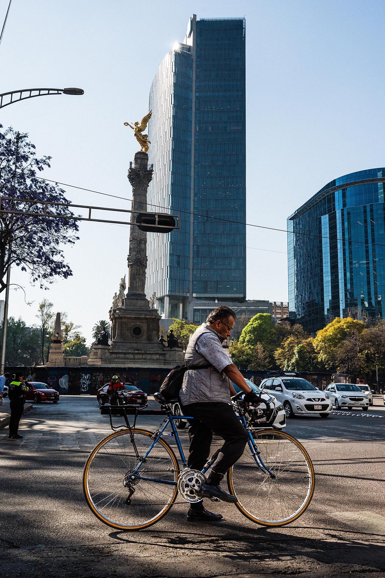 Cyclist rides past the Angel of Independence monument with modern skyscrapers in Mexico City's urban landscape.