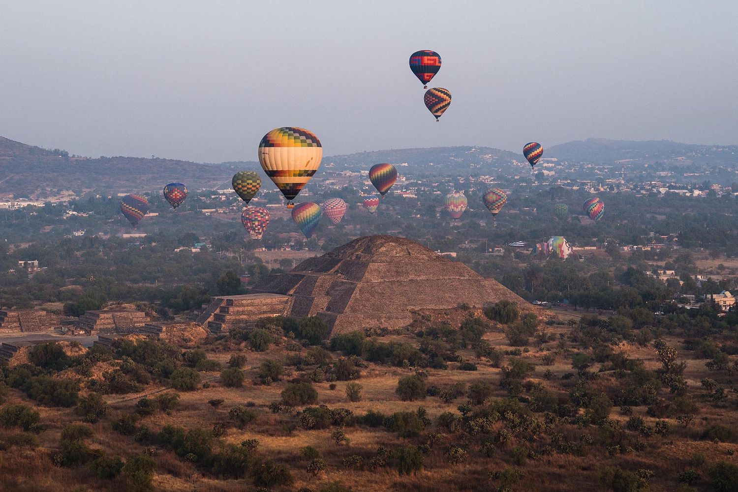 Hot air balloons float over a desert landscape during sunrise with mountains visible in the background.