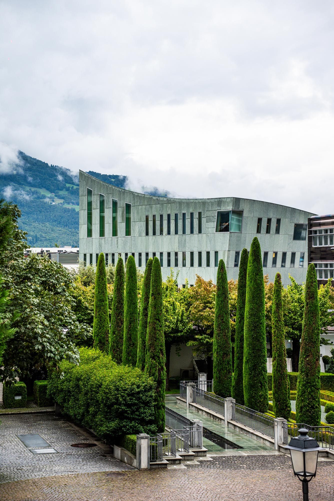 Modern architectural building with cypress trees lining the entrance pathway against a mountainous backdrop in cloudy weather.