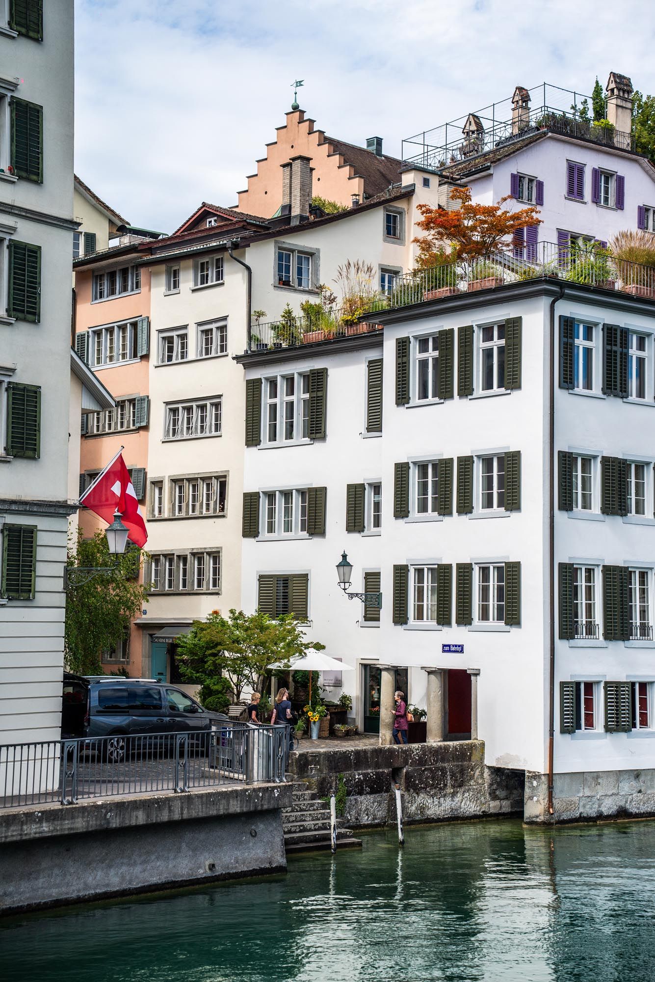 Traditional white buildings with Swiss flag along a canal in Zurich's old town district on a cloudy day.
