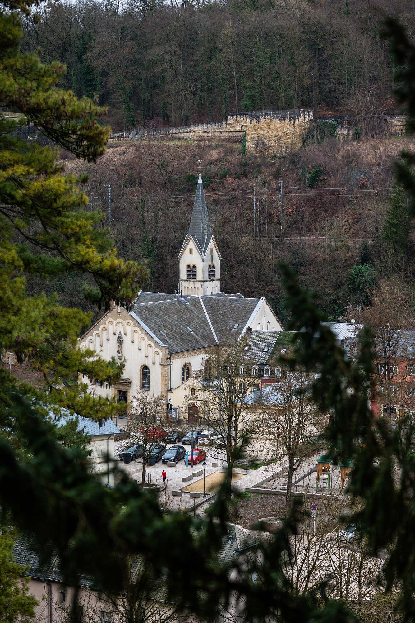 A white church with a steeple nestled among trees in a rural hillside setting viewed through pine branches.
