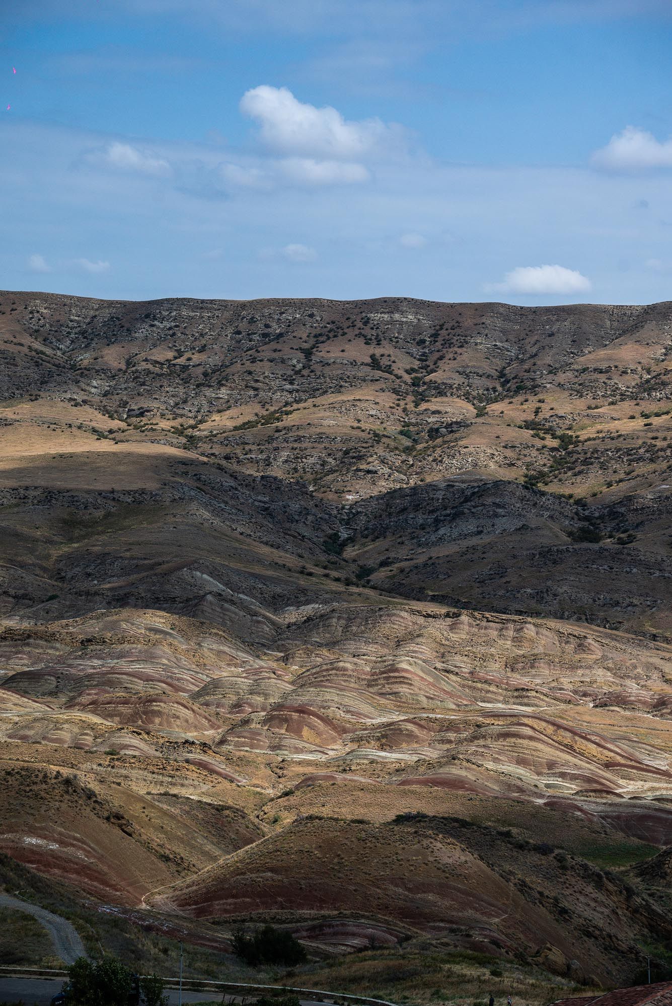 Layered sedimentary rock formations create colorful striations across an arid mountainous landscape under blue skies.
