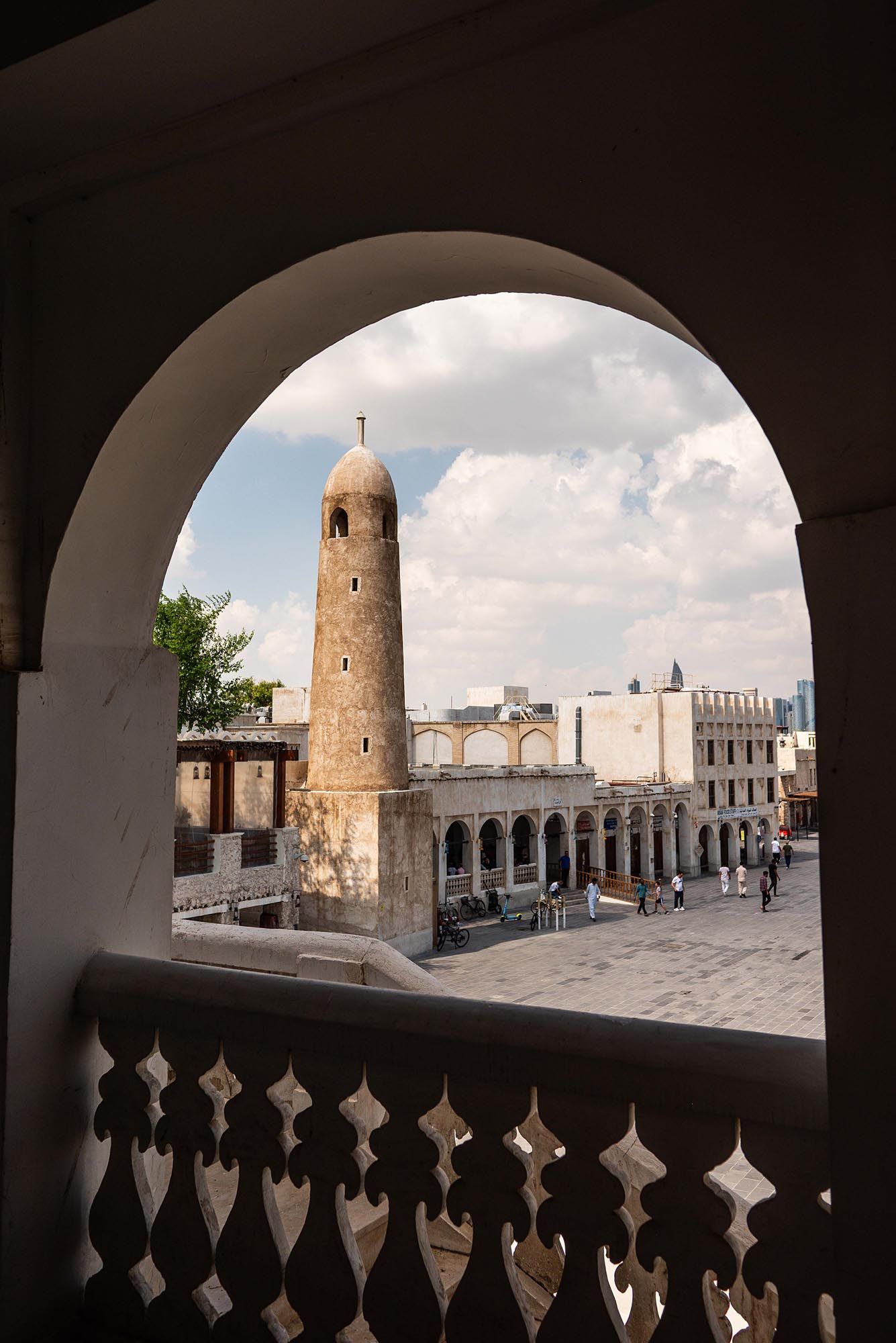 View through ornate arches of a historic mosque courtyard with minaret tower and white buildings in Qatar.