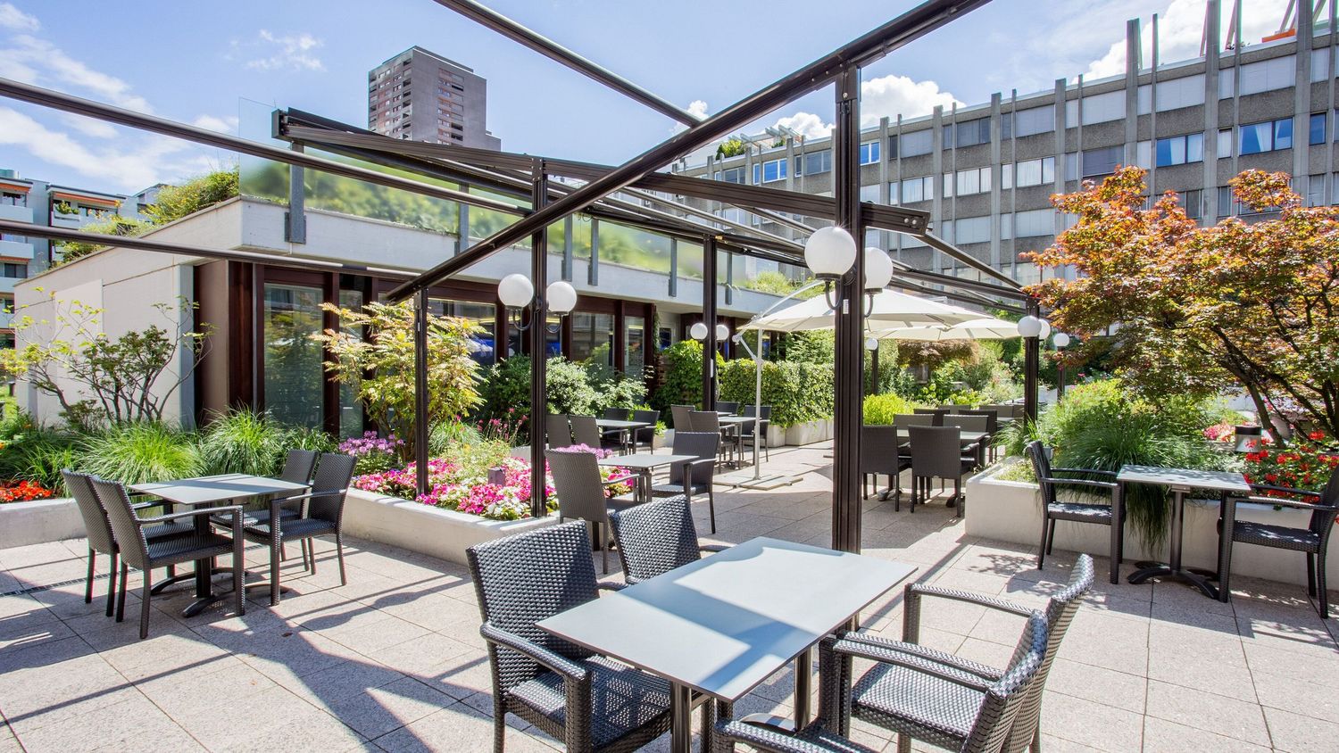 Modern rooftop terrace with comfortable seating and glass railings overlooking a city skyline on a sunny day.