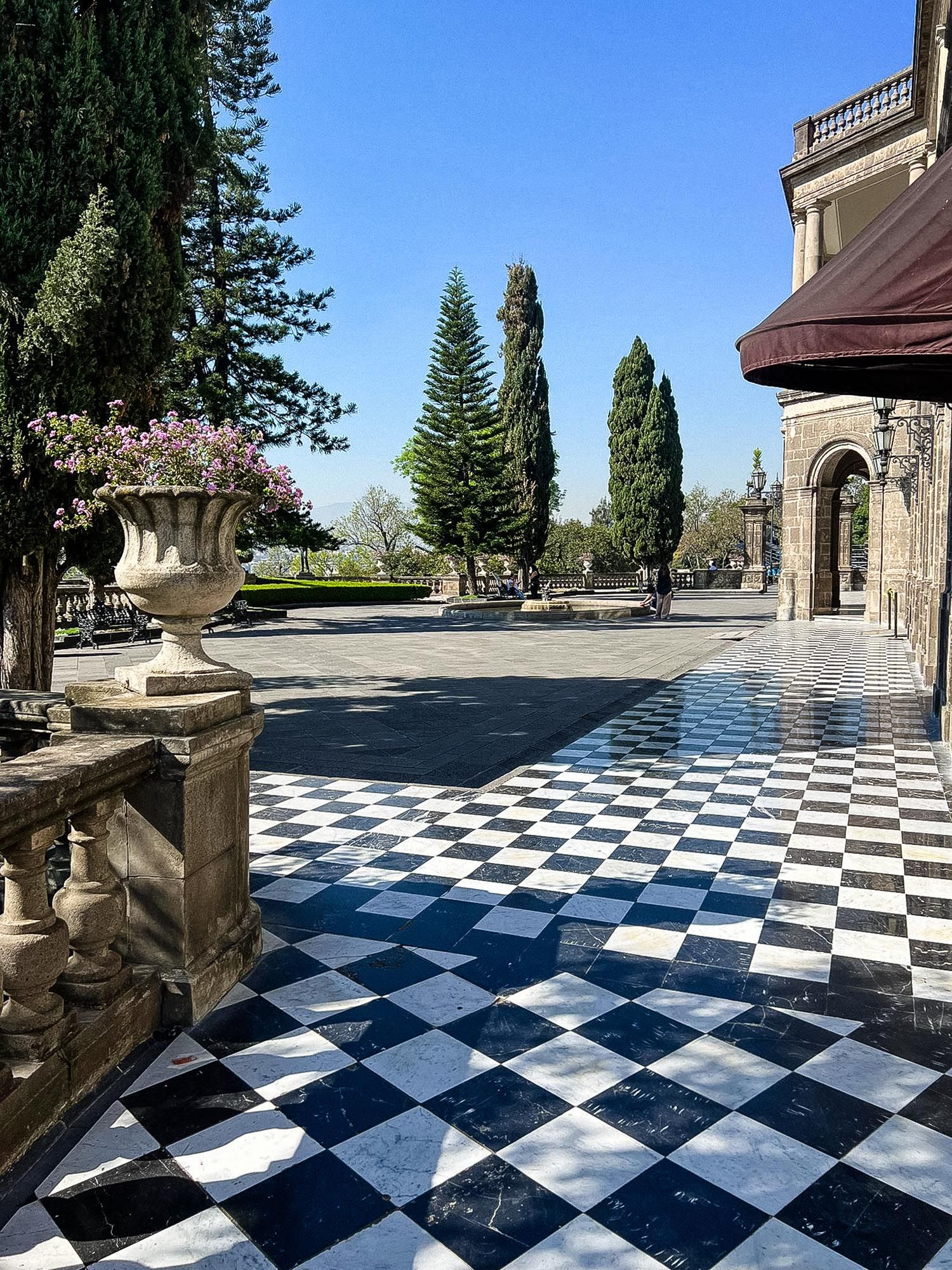 Black and white checkered marble terrace with stone balustrades and evergreen trees creates a dramatic outdoor space.