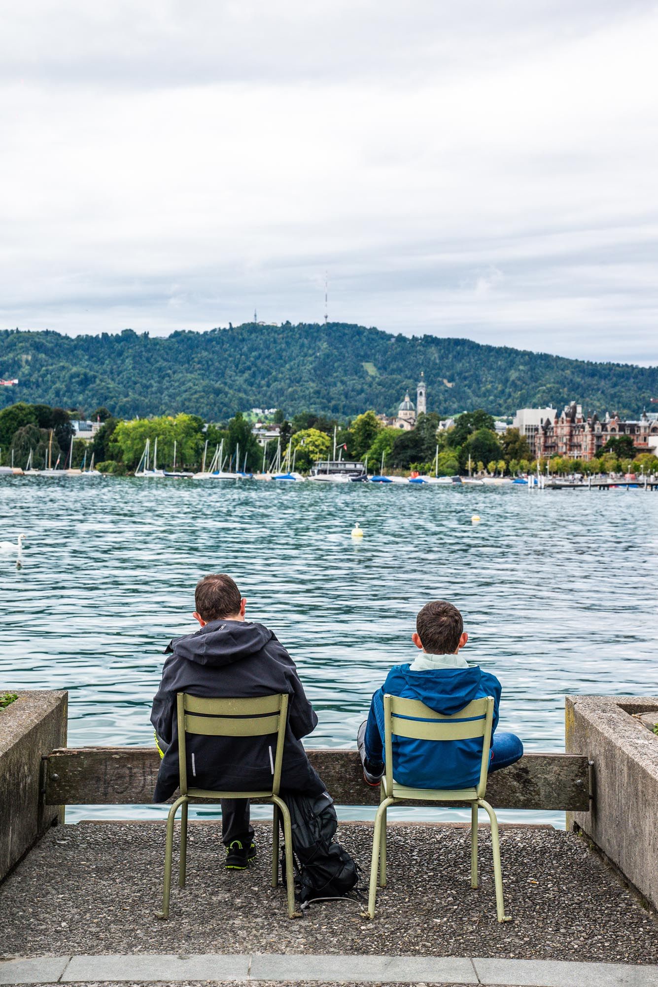 Three people sit in chairs along Lake Zurich's waterfront, gazing at boats and forested hills on a cloudy day.