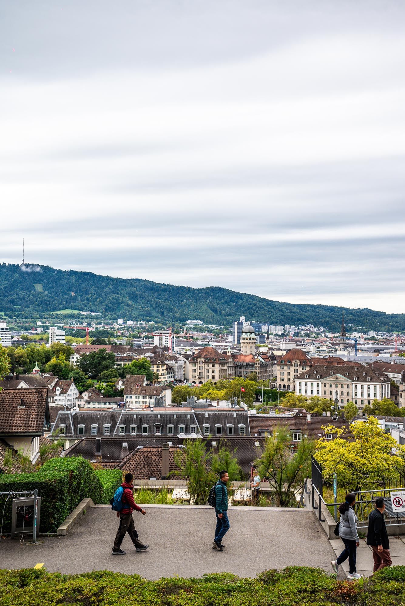 People enjoy panoramic views of Zurich from a scenic overlook with mountains and city buildings in the background.