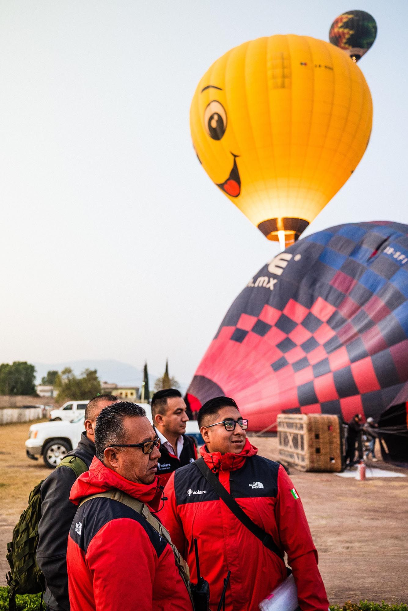 Hot air balloon crews prepare for launch at dawn as a yellow smiley face balloon rises in the background.