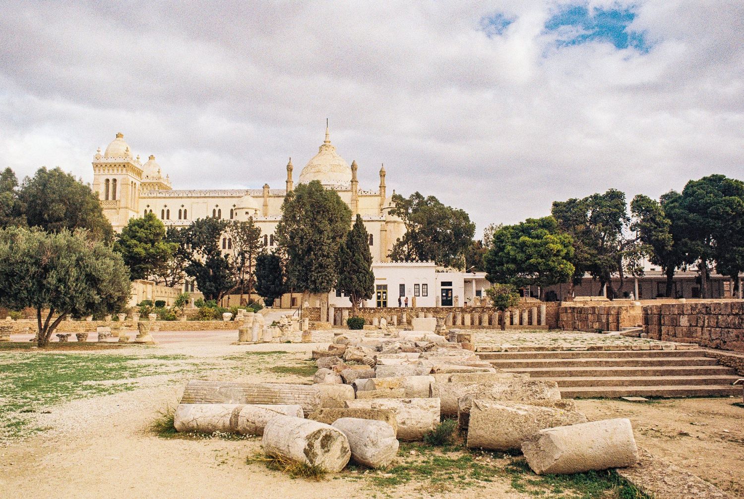 Ancient Roman ruins with fallen columns in foreground and historic buildings against cloudy sky in Malta.