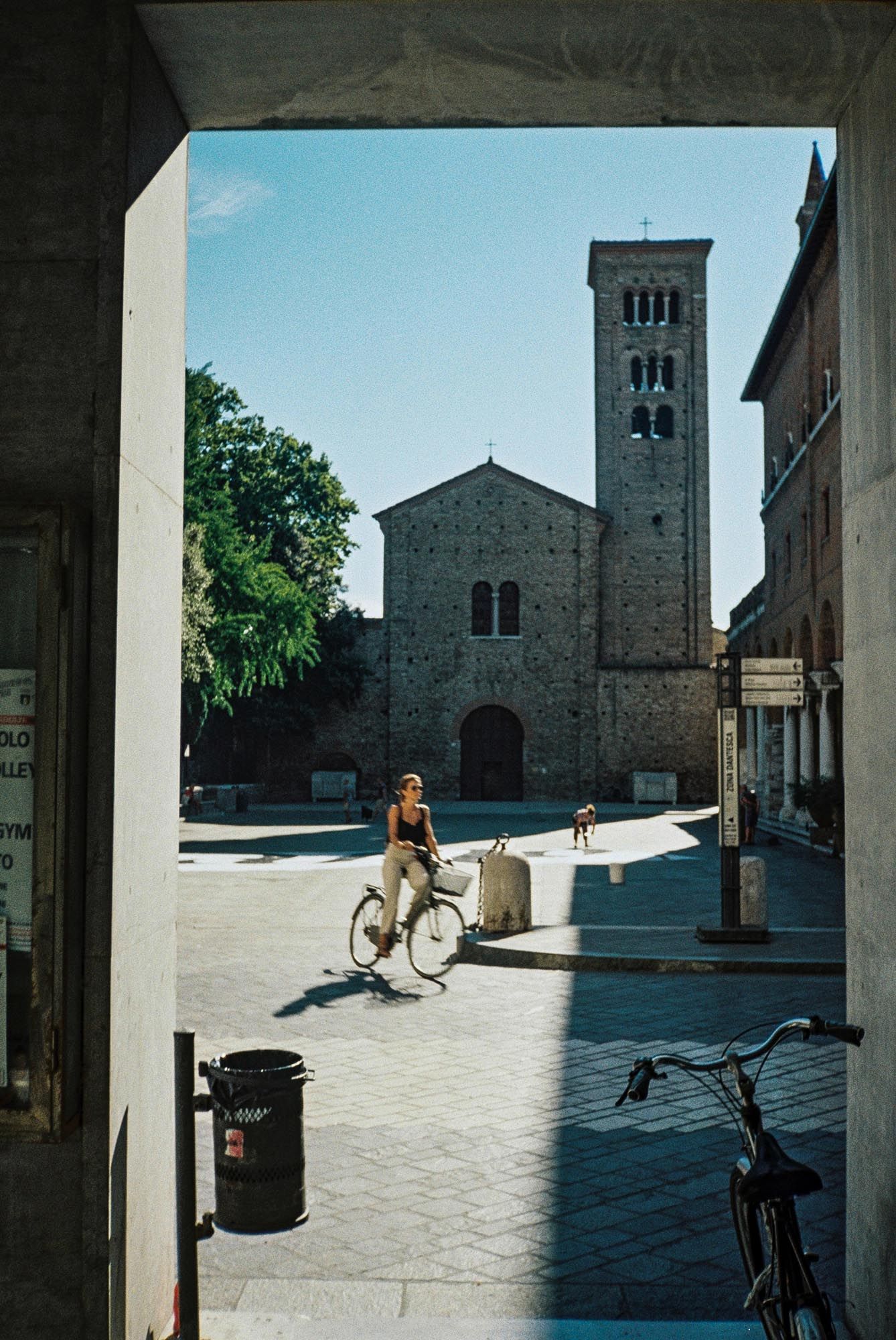 A panoramic view of a historic Italian piazza with stone buildings, church facade, and bicycles in the sunlit square.