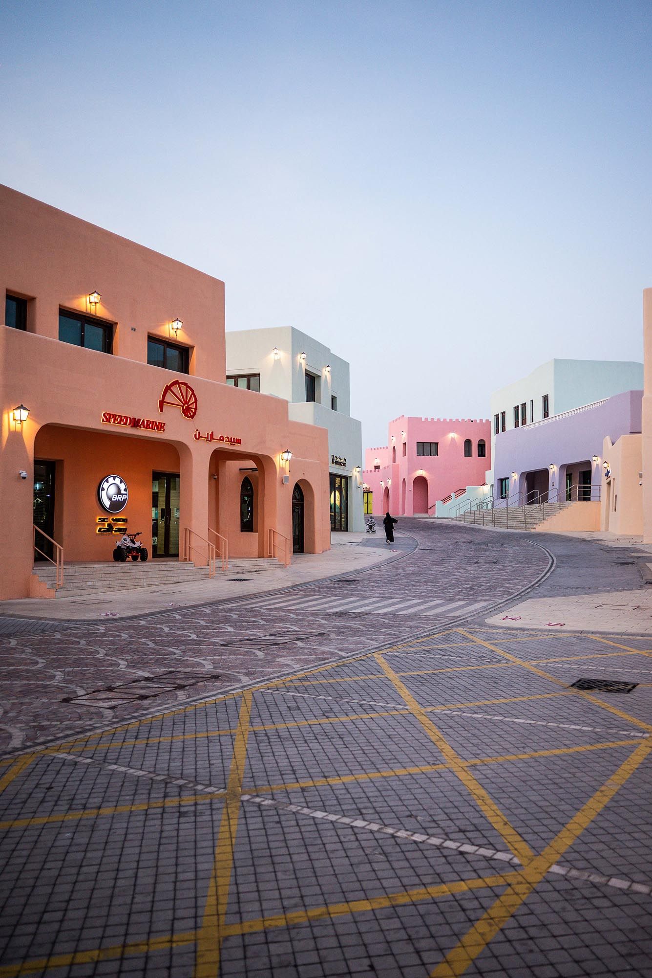 Curved street lined with modern pastel-colored buildings and geometric parking spaces in a desert setting at dusk.