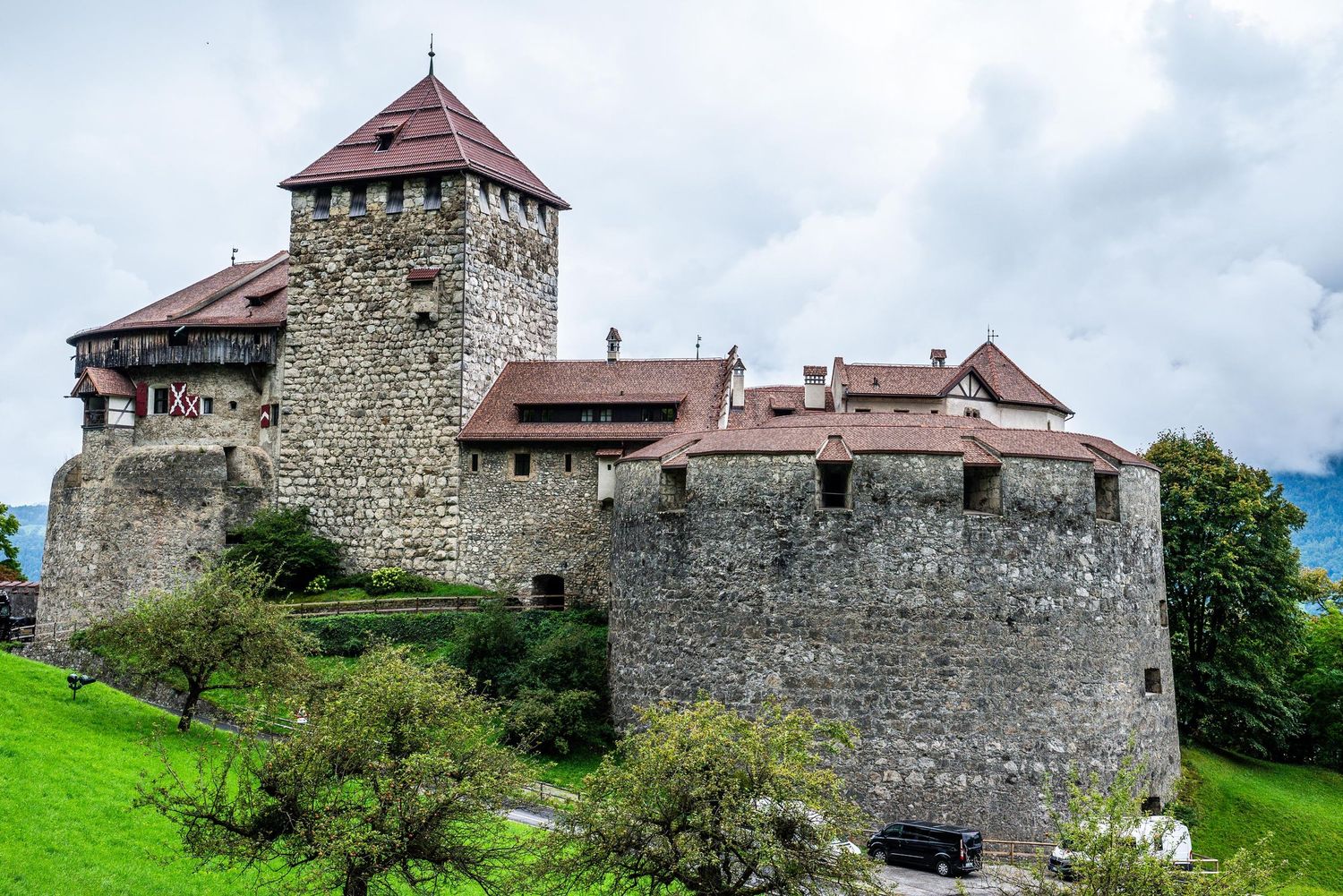 Medieval stone fortress with red-tiled towers sits atop a green hillside surrounded by trees and countryside.