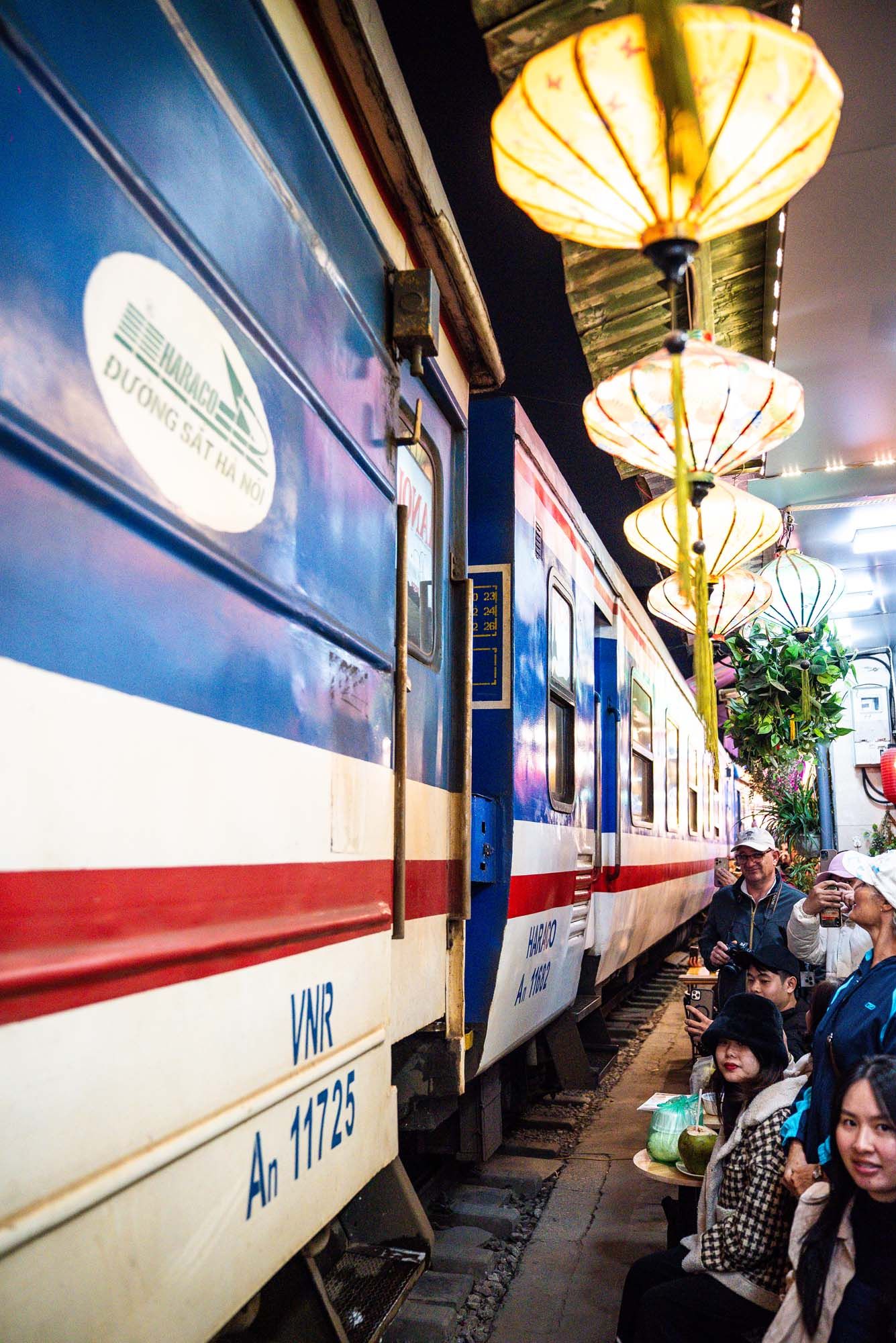 Passengers board a white and red striped train at a dimly lit station platform with ornate lantern lighting.