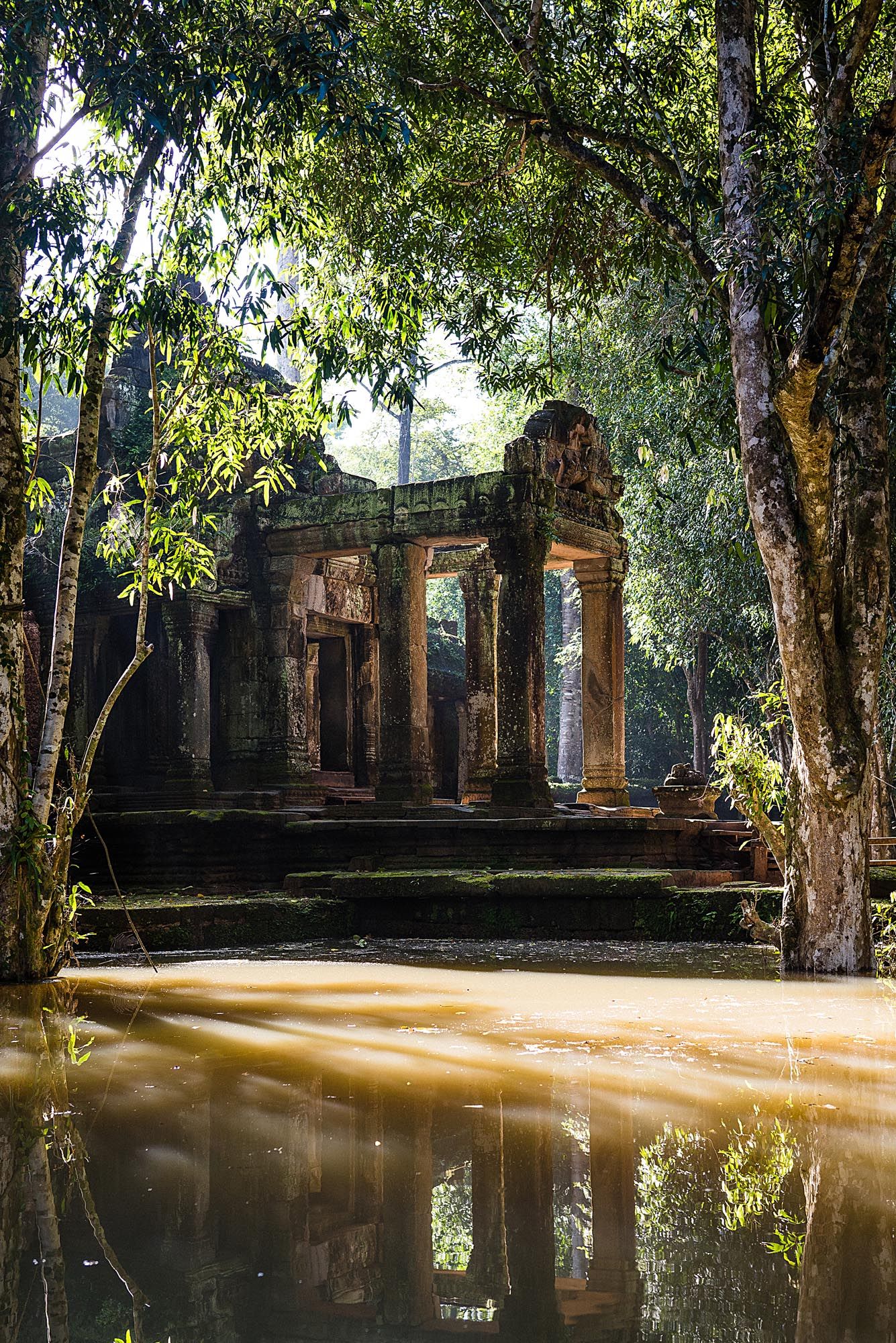 Ancient temple ruins surrounded by trees reflect in still floodwaters at a historic archaeological site in Southeast Asia.