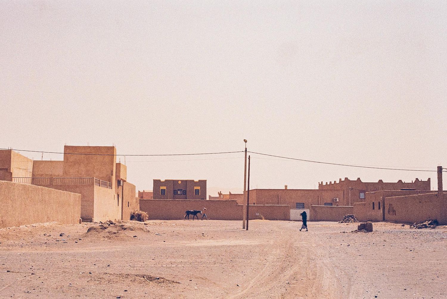A lone figure walks down a dusty road between clay buildings in a desert town with utility poles.