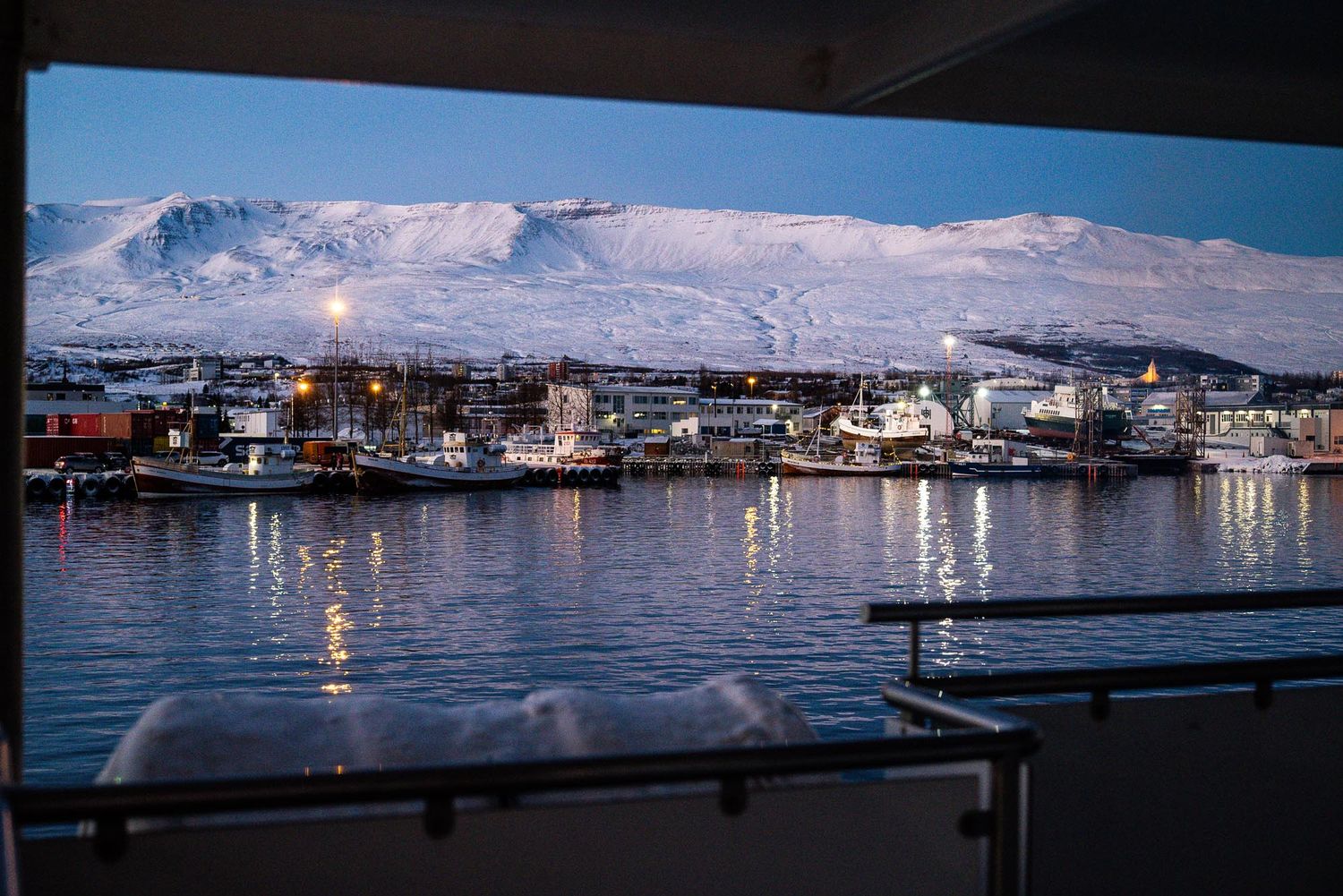 Panoramic view of boats docked in a harbor at dusk with city lights reflecting on calm waters.