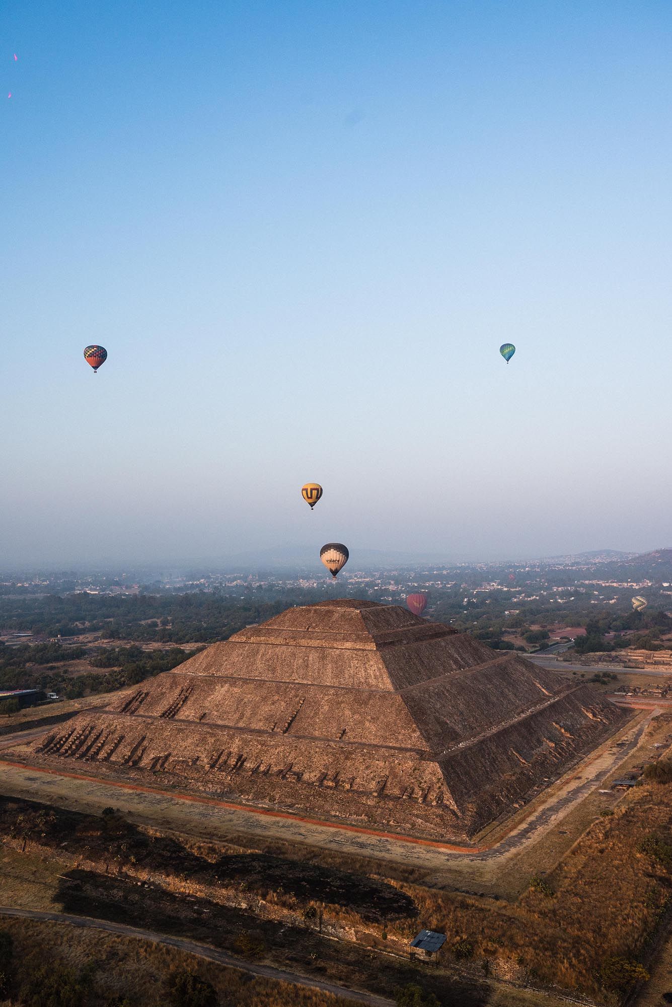 Hot air balloons float above an ancient pyramid-shaped structure against a clear blue sky in a desert landscape.