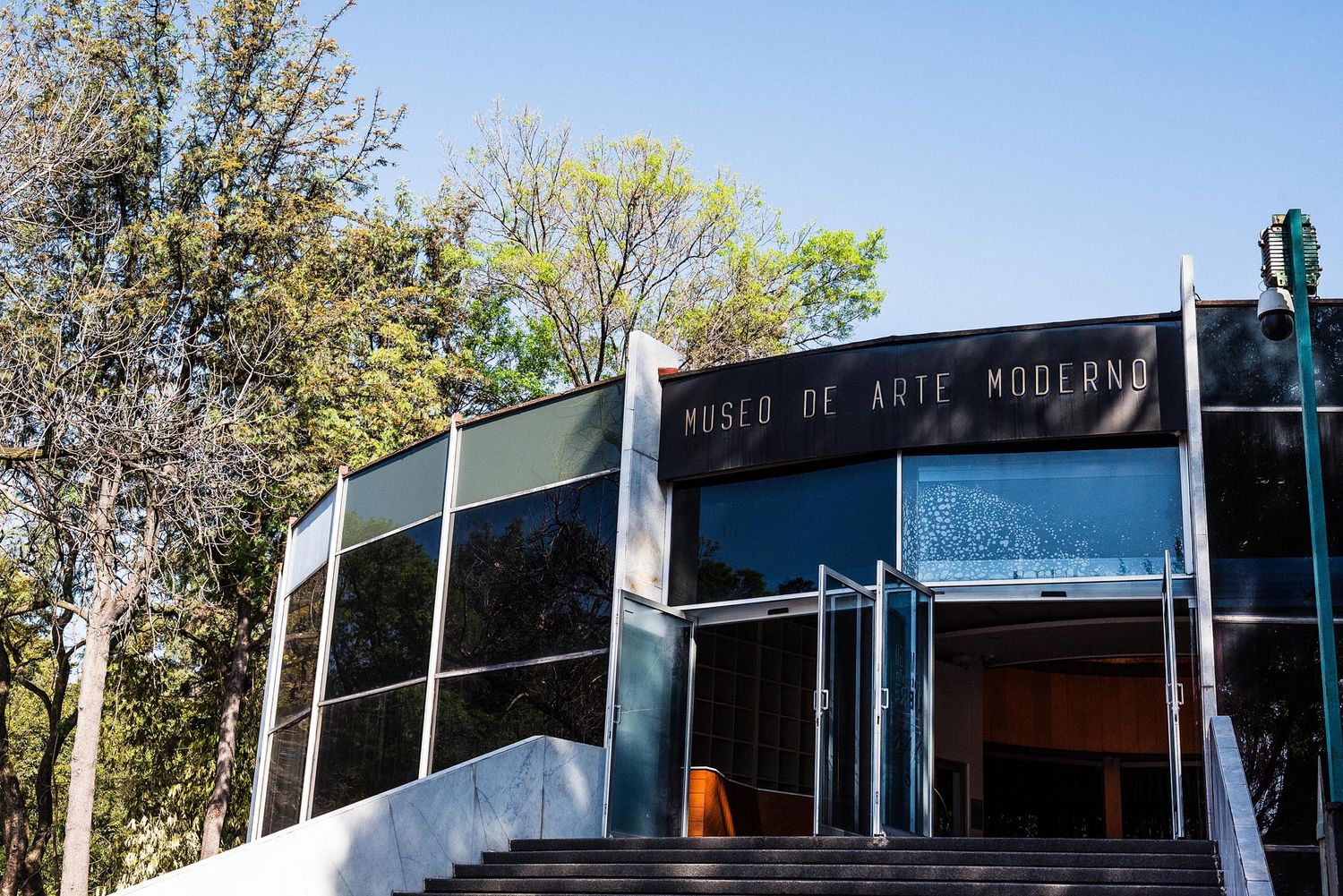Modern glass and steel entrance to the Museum of Modern Art building with trees visible in background.