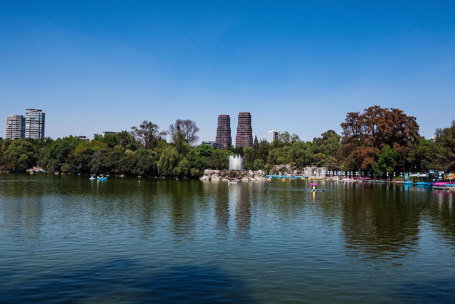 Panoramic view of a serene lake surrounded by trees and city buildings under a bright blue sky.