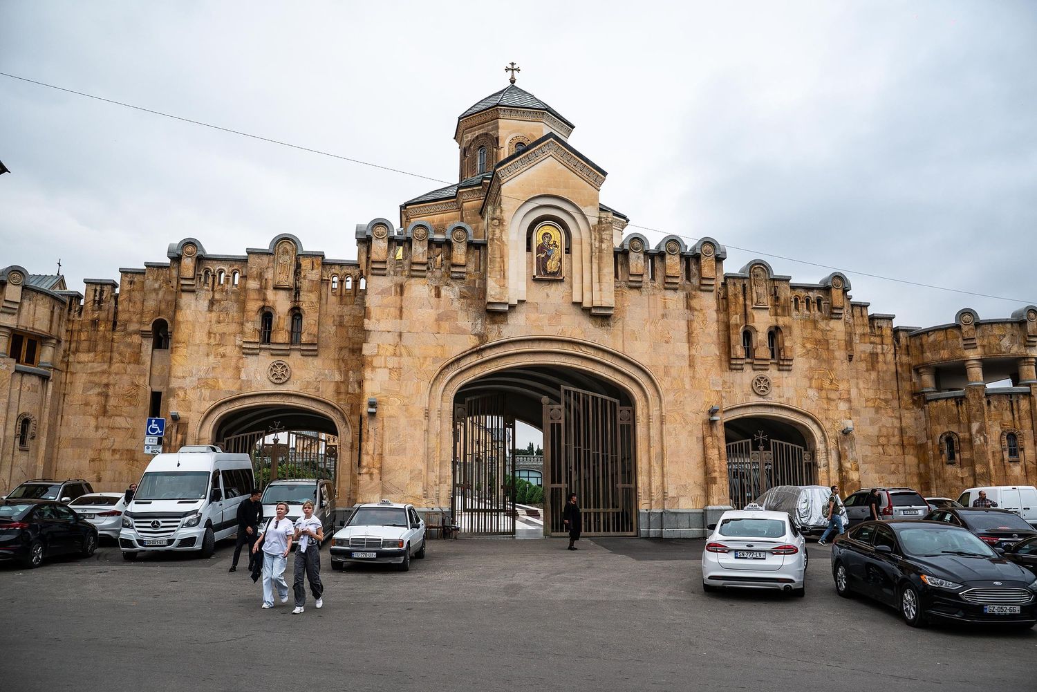 Historic stone walls and arched entrances of Echmiadzin Cathedral in Armenia with parked cars in the foreground.