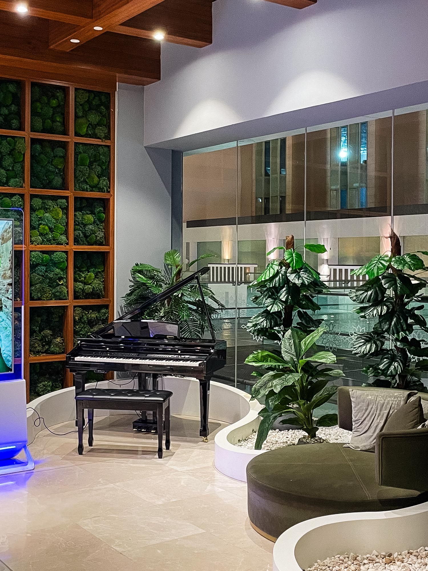 Modern office lounge area with curved seating, indoor plants, and wooden beam ceiling accents illuminated by ambient lighting.