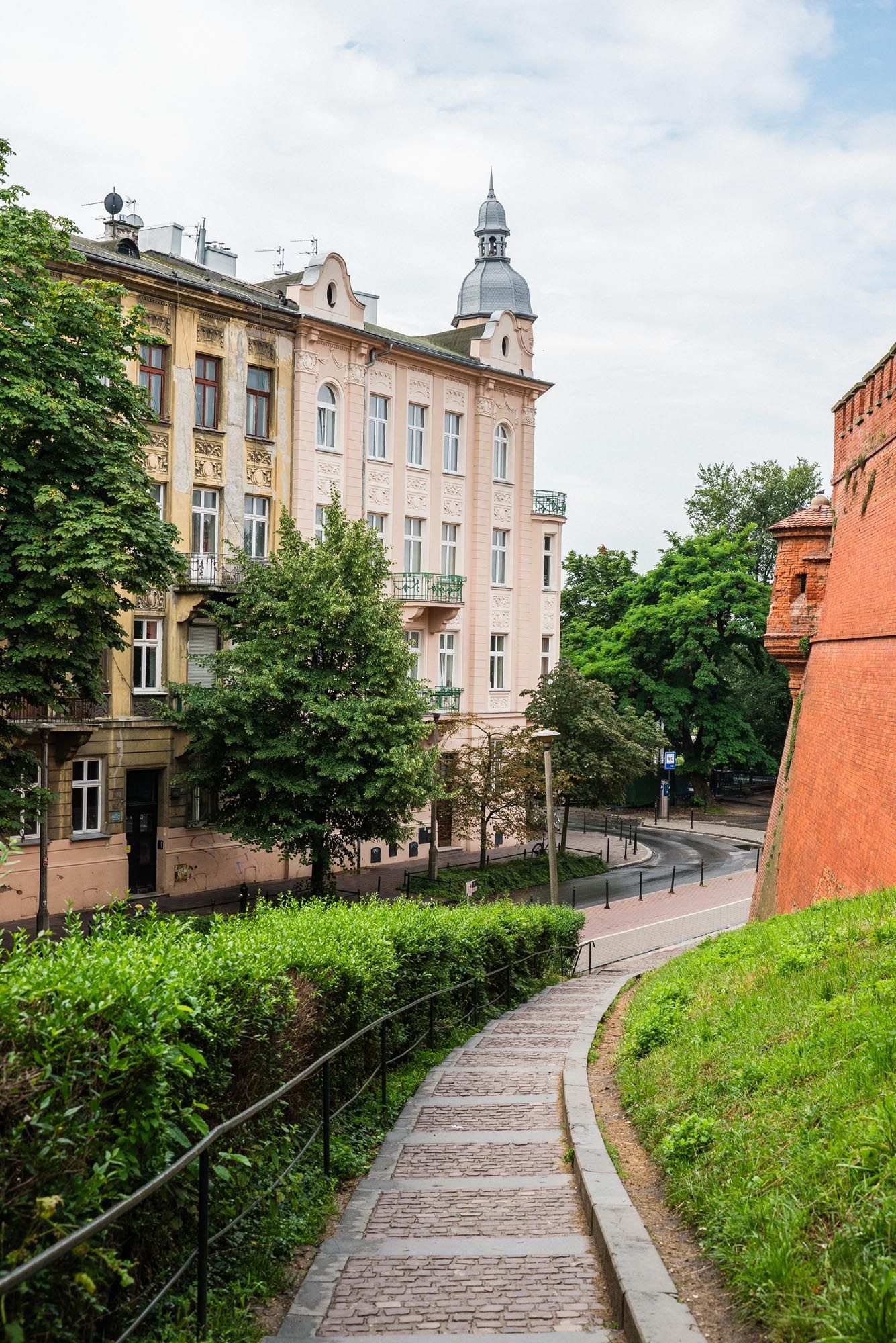 Curved stone pathway leads up a green hill past pink historic building with tower dome in Krakow, Poland.