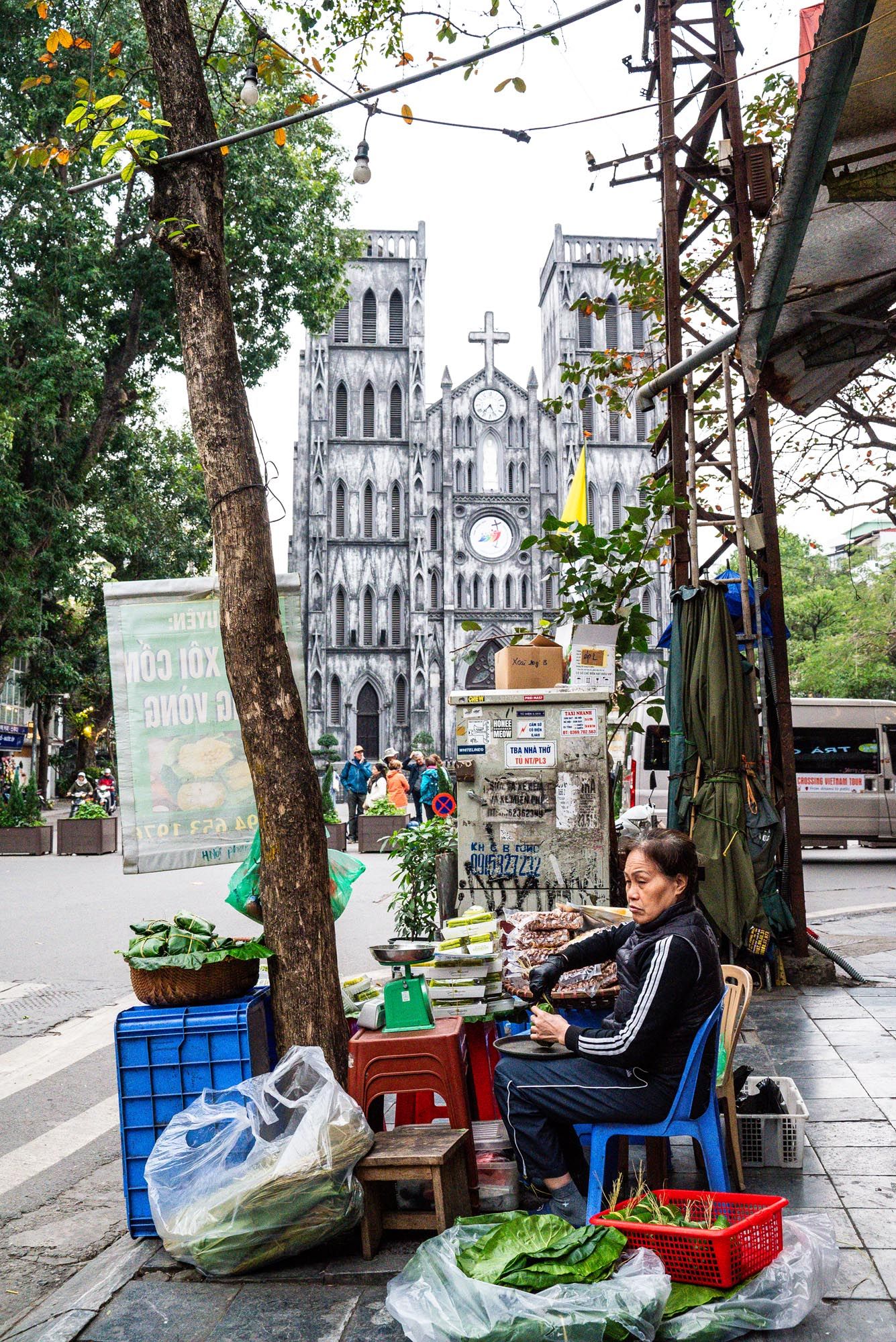 Street vendor sells fresh produce outside Saint Joseph's Cathedral in Hanoi's Old Quarter with Gothic towers in background.