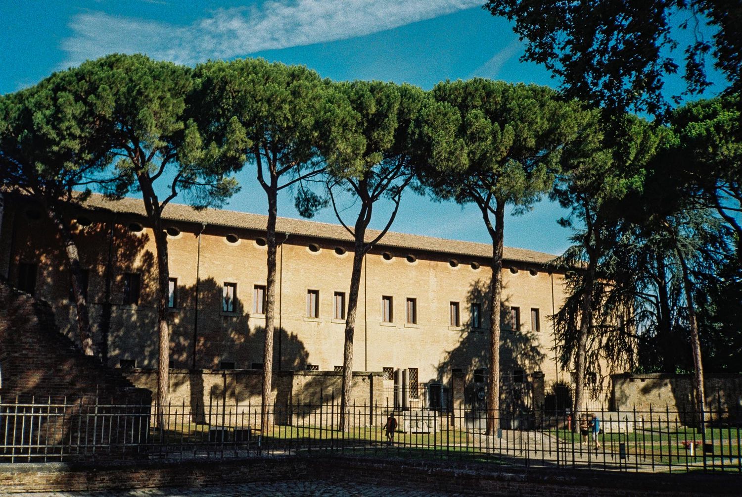 A panoramic view of a beige stone building complex with Mediterranean architecture nestled among green trees under blue skies.