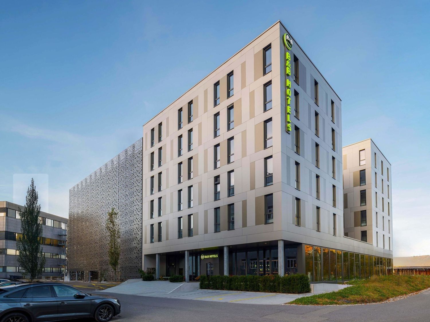 Modern multi-story hotel building with beige exterior walls and vertical green signage against a blue sky.