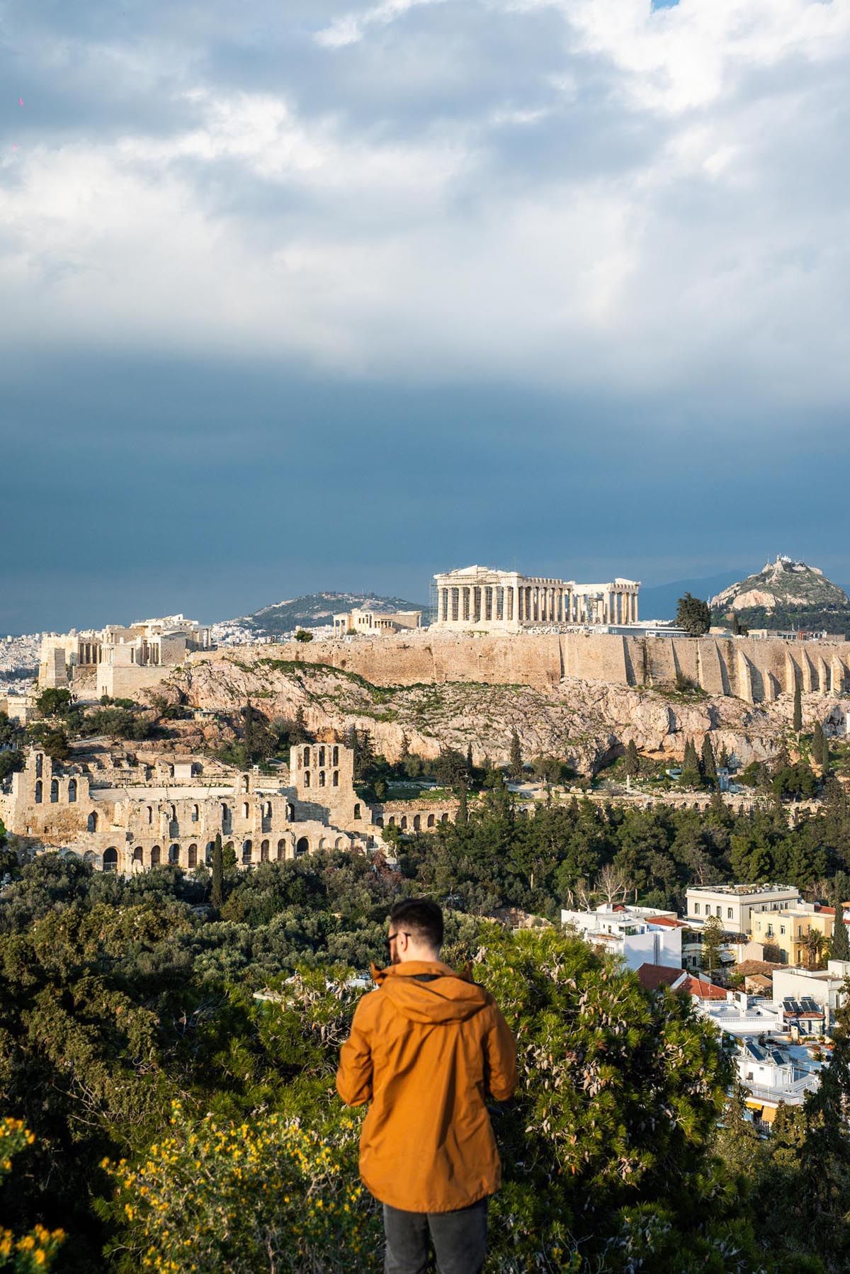 Panoramic view of the Acropolis and Parthenon in Athens with a person in orange jacket viewing the ancient ruins.