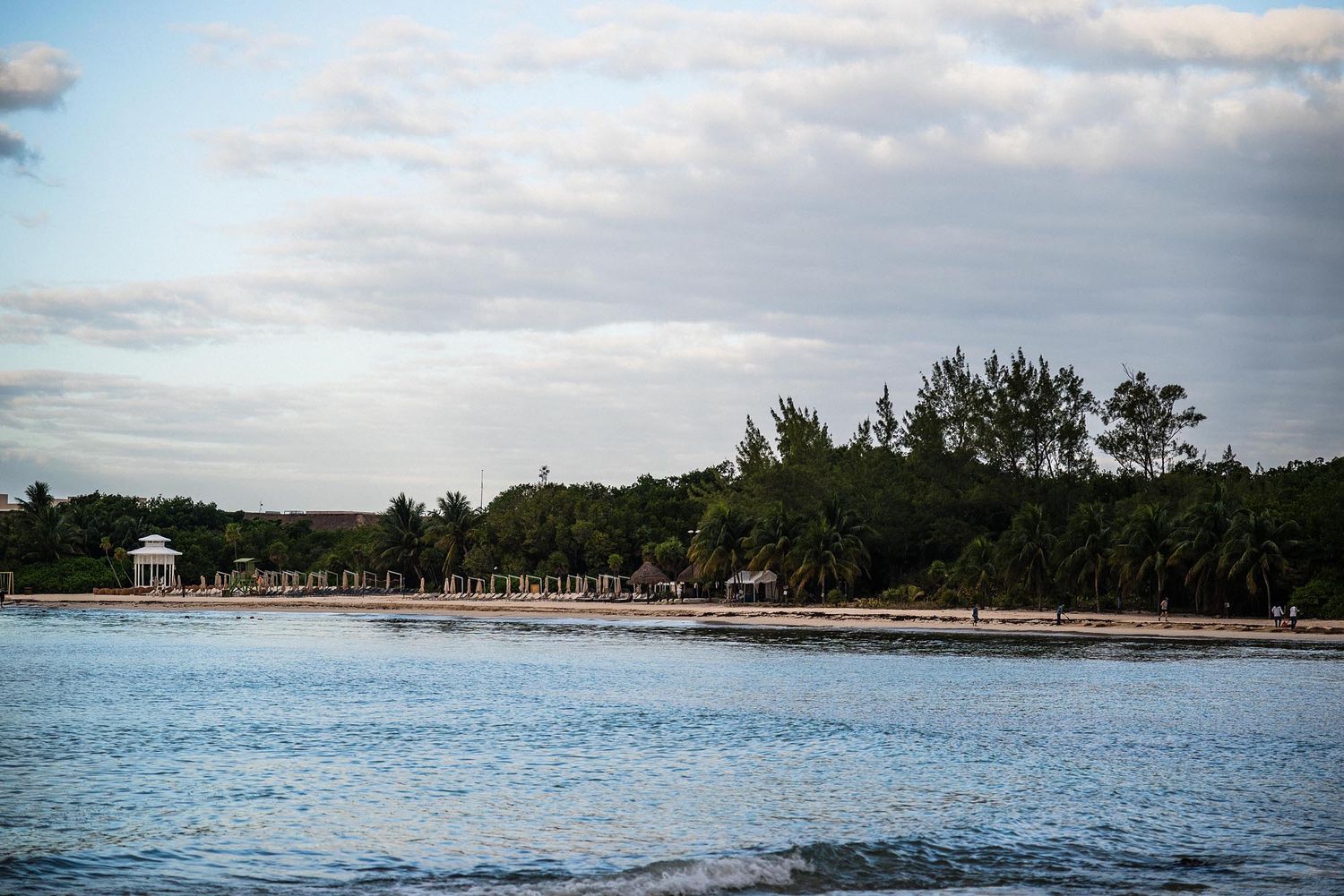 Tranquil beach shoreline with turquoise waters and tree-lined coast under cloudy skies in tropical setting.