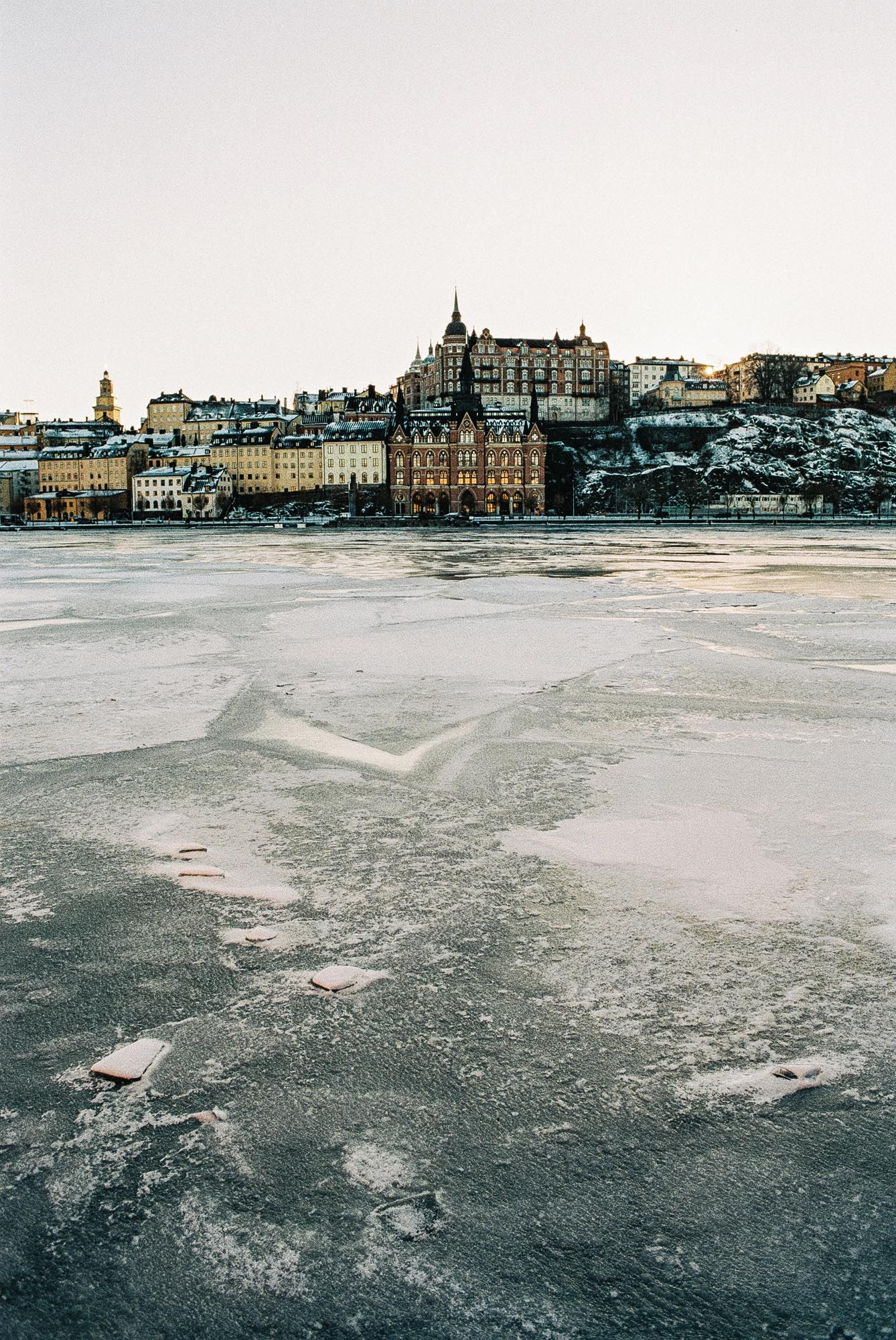 A panoramic winter view of historic buildings along a frozen lake shore with snowy ice formations in the foreground.