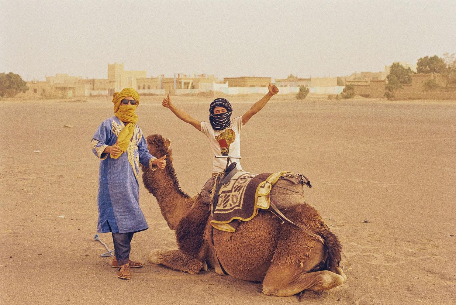 A group of tourists ride camels across a vast desert landscape at sunset.