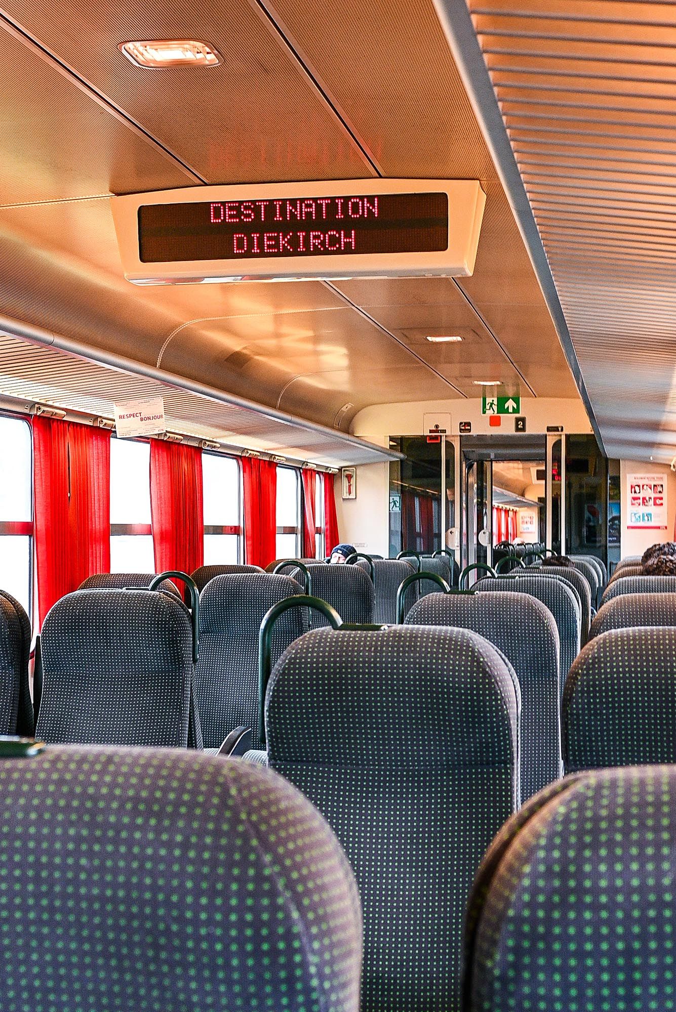 Empty passenger seats inside a modern train car with blue upholstery and a digital destination display showing Biekirch.