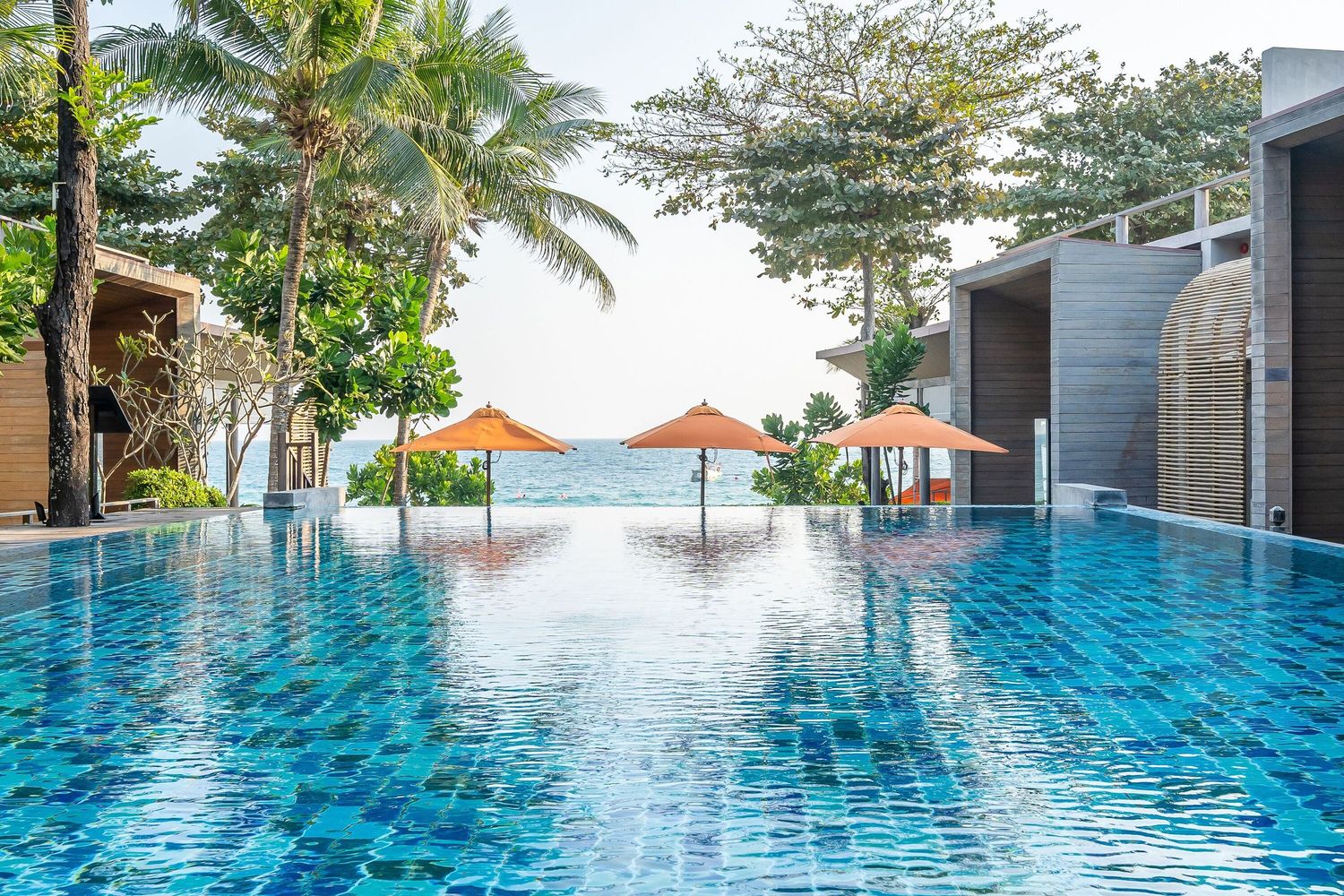 Luxury infinity pool overlooking tropical ocean with palm trees and orange umbrellas at sunset.