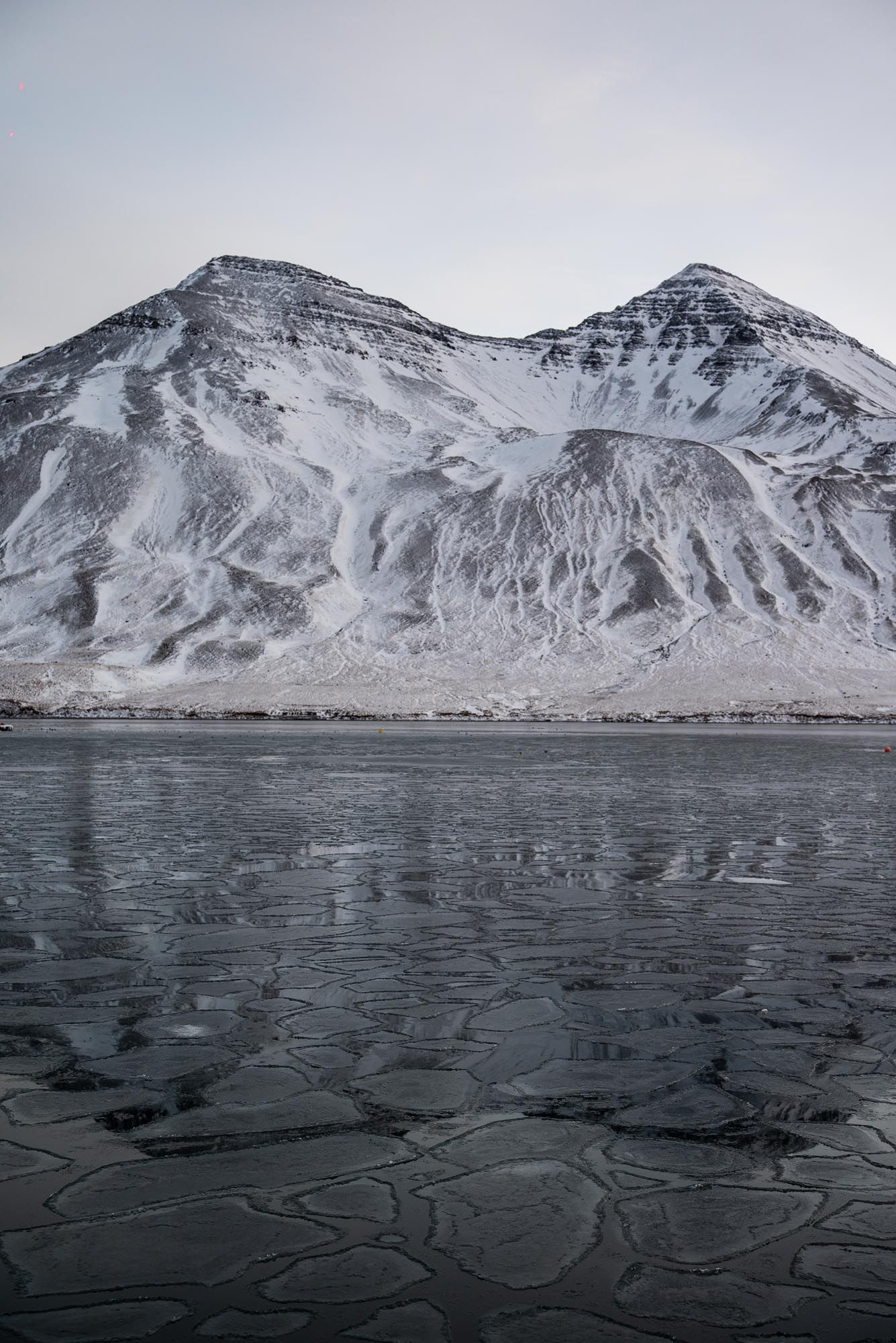 Snow-covered mountain peaks rise above a dark frozen lake with intricate cracking patterns on the icy surface.
