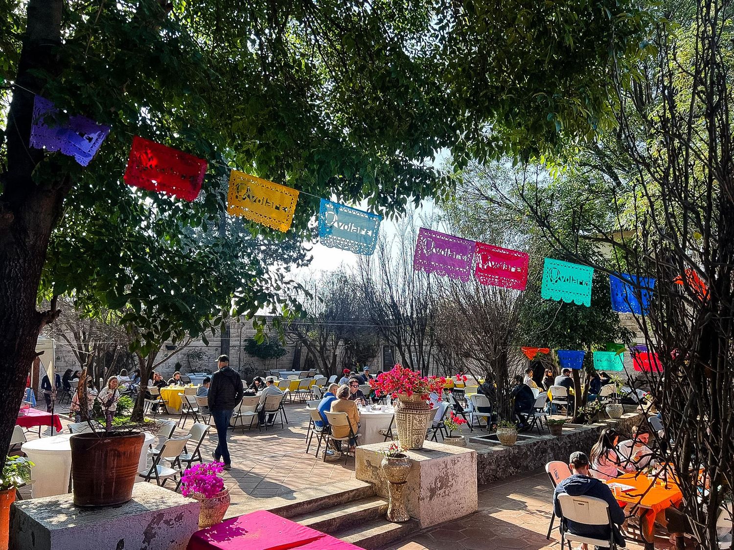 Colorful papel picado banners hang over an outdoor market area with vendors and trees providing shade.