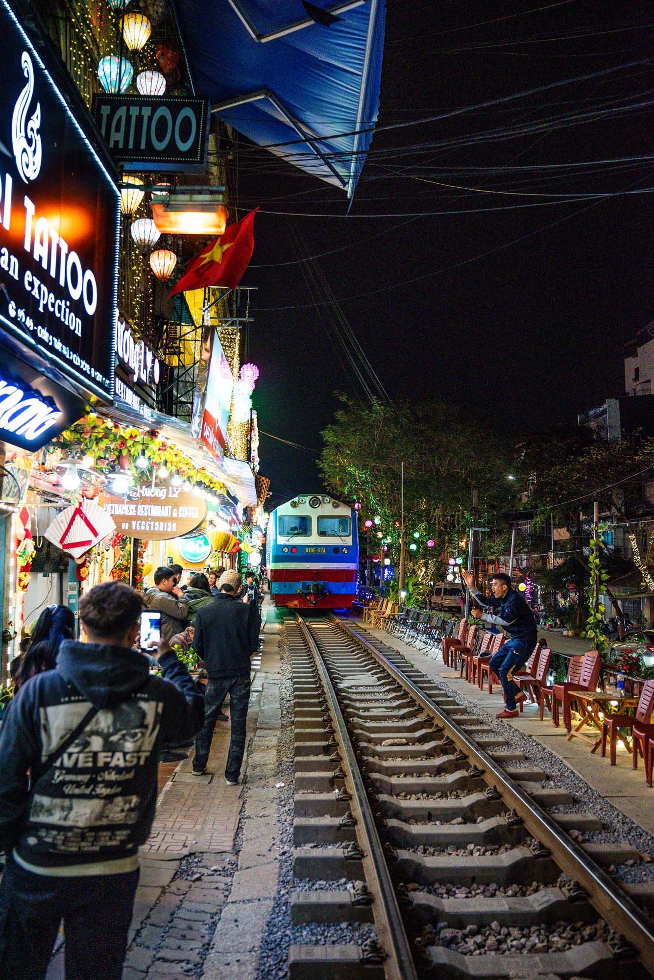 Train tracks run through a bustling night market in Hanoi with glowing lanterns and shops along both sides.
