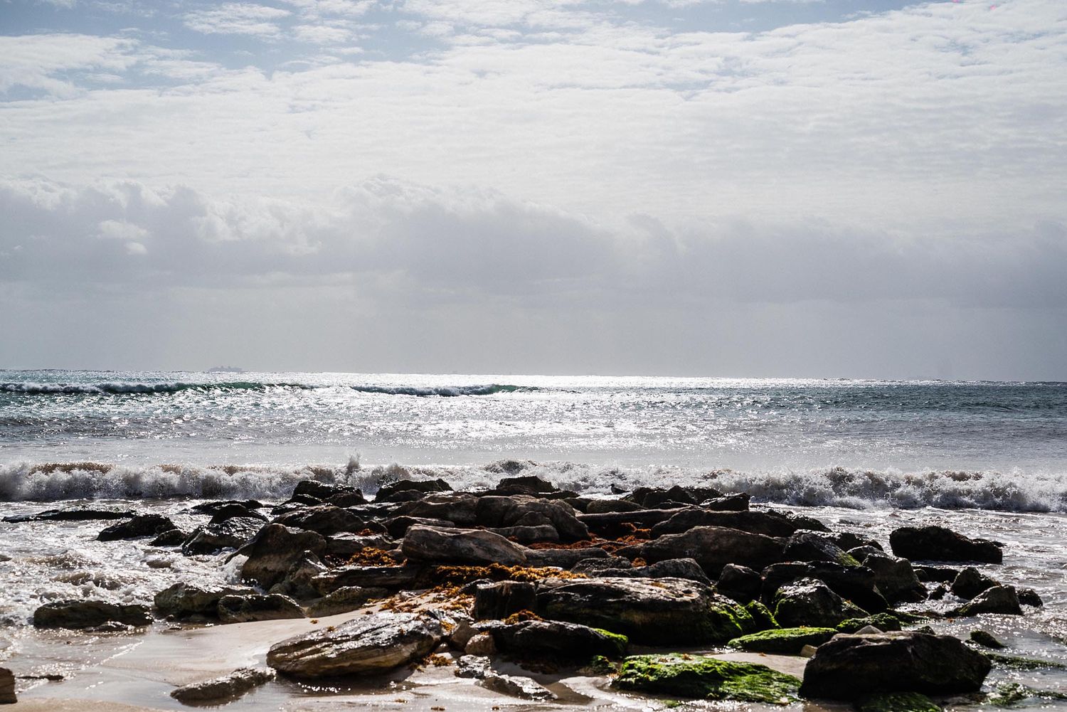 Panoramic view of rocky coastline with crashing ocean waves under cloudy sky and sparkling sunlight on water.
