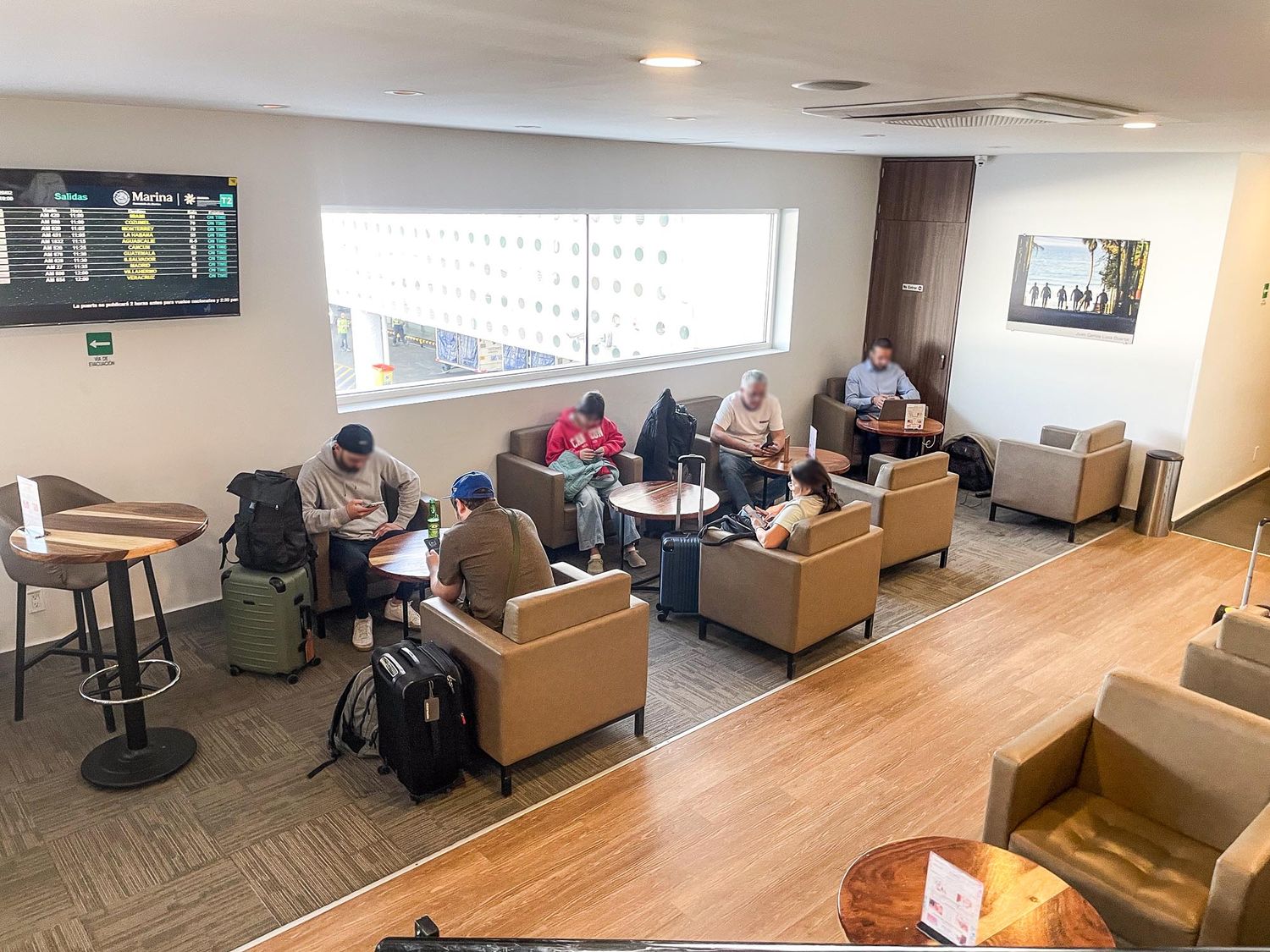 Modern airport lounge area with tan couches, wood flooring, and digital departure board displays mounted on walls.