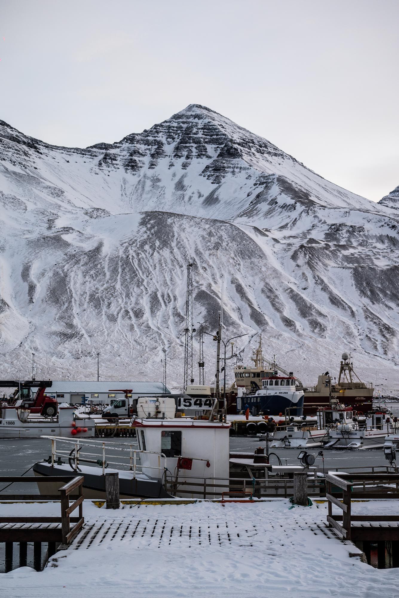 Snow-covered mountains tower over a harbor filled with fishing boats and industrial vessels in a winter landscape.
