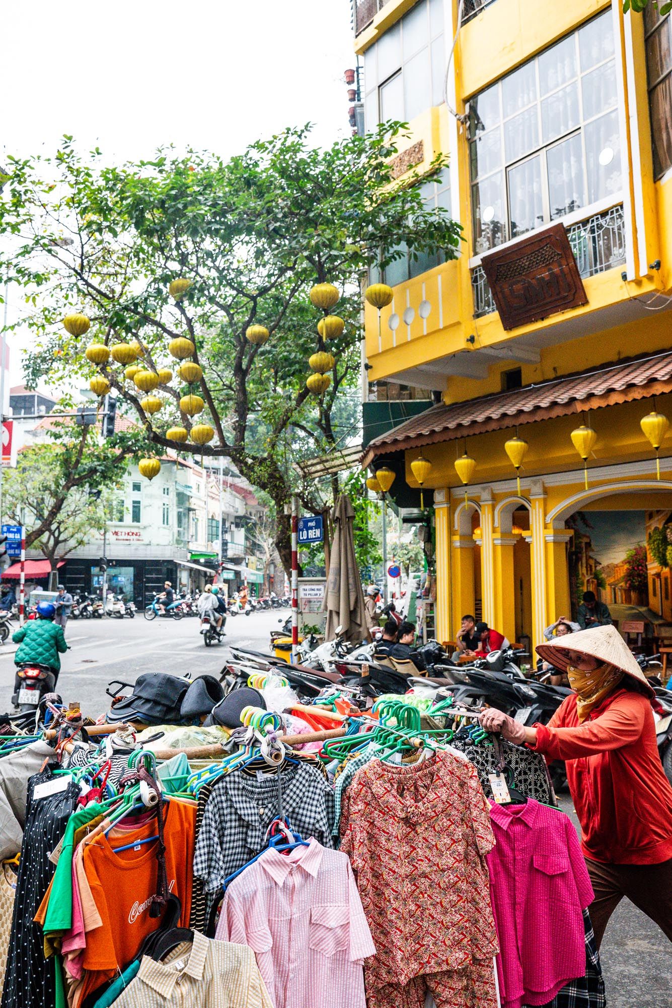Colorful clothing displayed at street market stall with yellow lanterns and building facade in Vietnam.