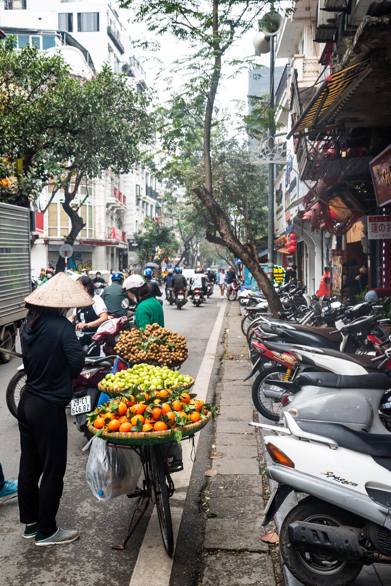 Street vendor selling fresh fruit from bicycle cart on busy street lined with scooters in Hanoi, Vietnam.