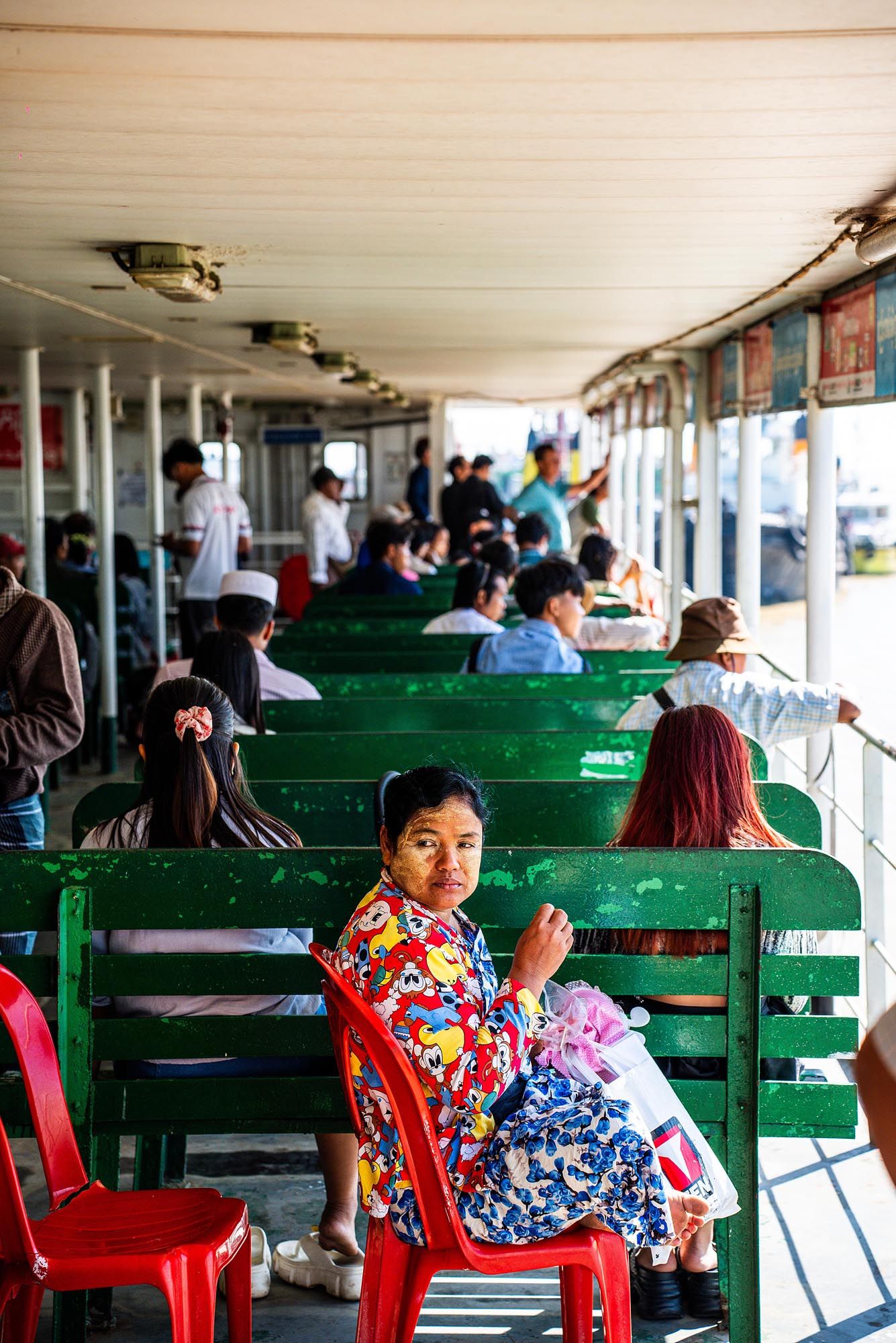 Passengers sit on green wooden benches inside a ferry boat cabin during a journey across water.