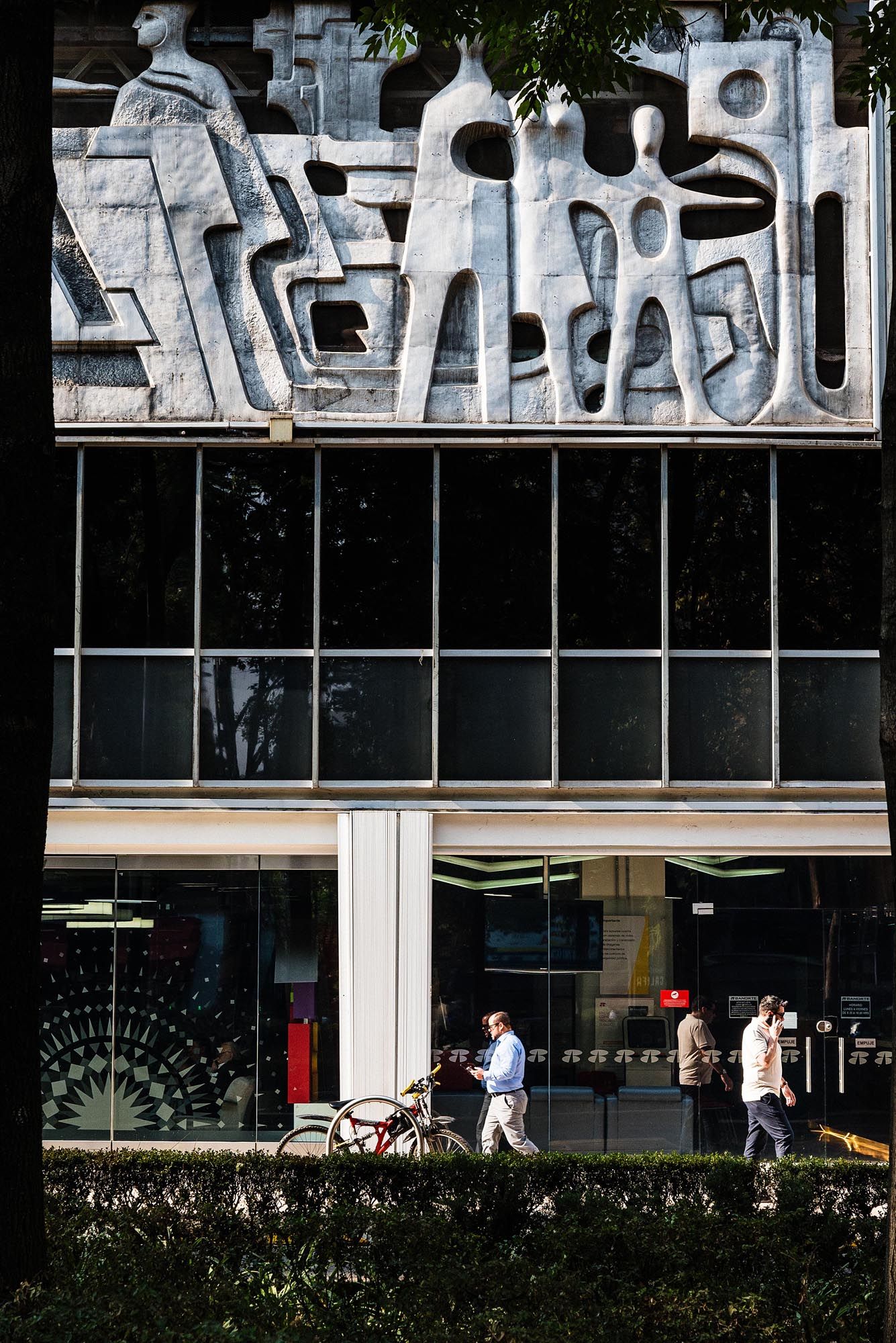 People walking past a modern building with geometric text signage and glass windows on a sunny day.