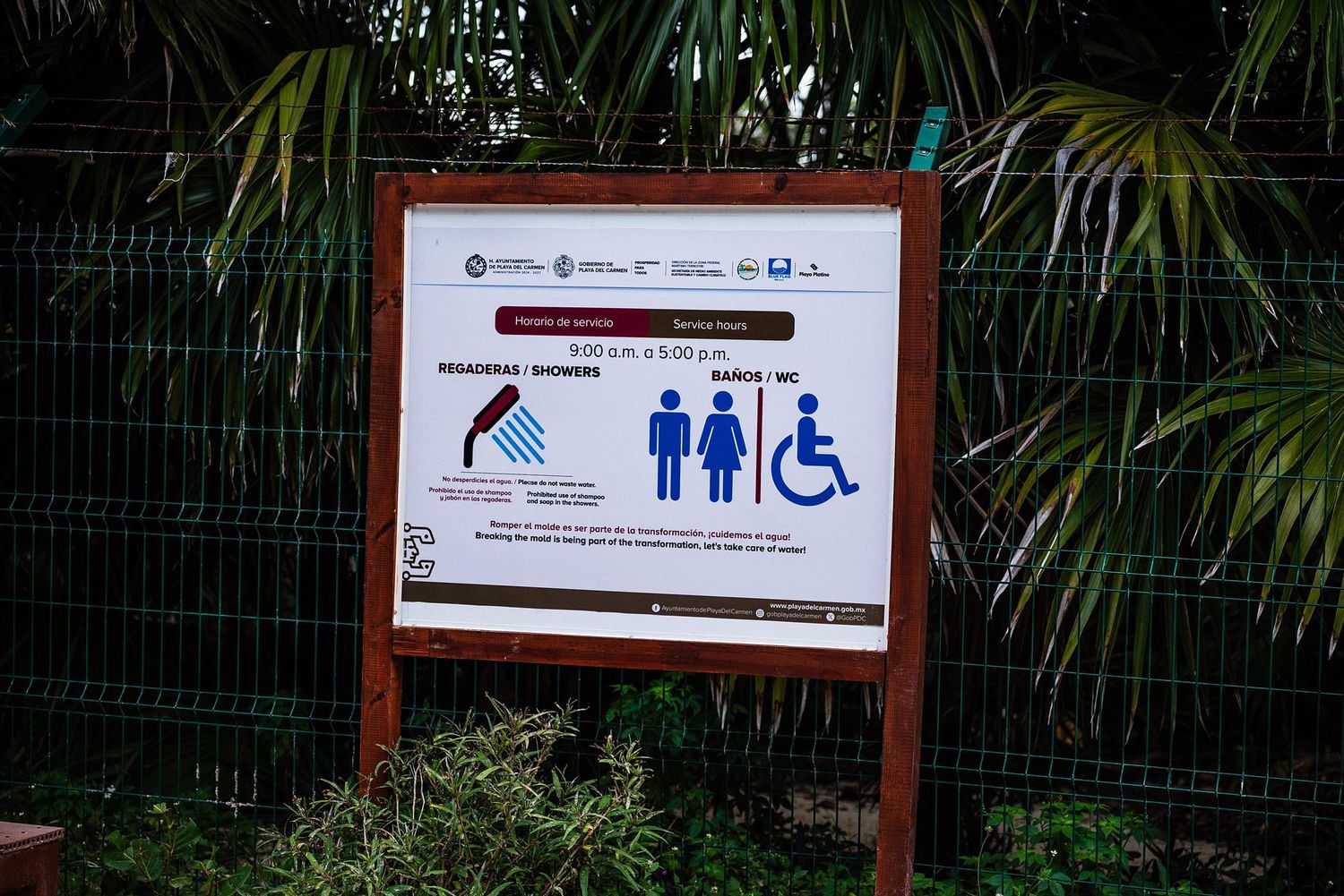 A wooden sign with bathroom symbols and accessibility icons mounted on a fence with tropical plants in the background.